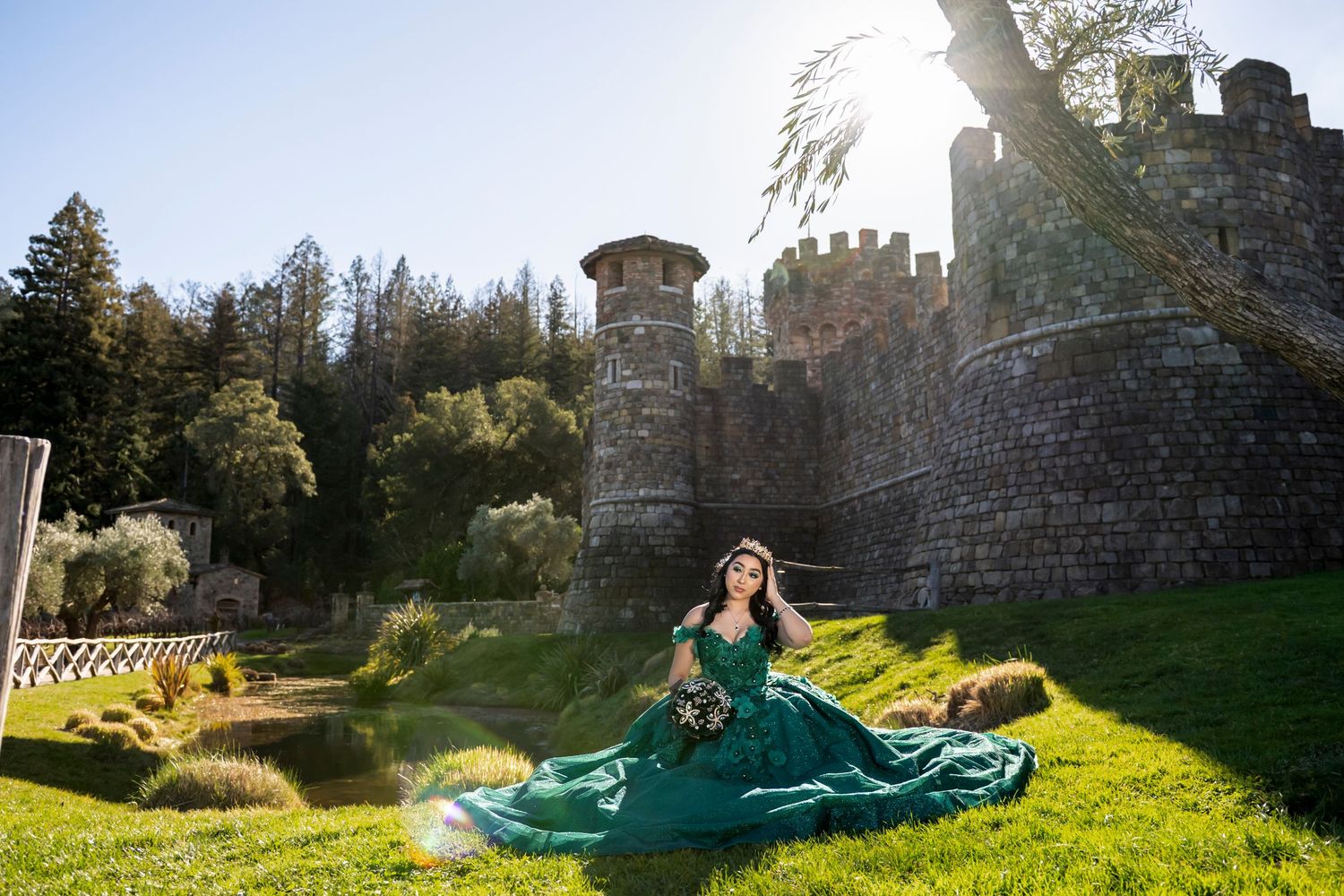 Woman in emerald green ball gown poses on grass before a stone castle on a sunny day.