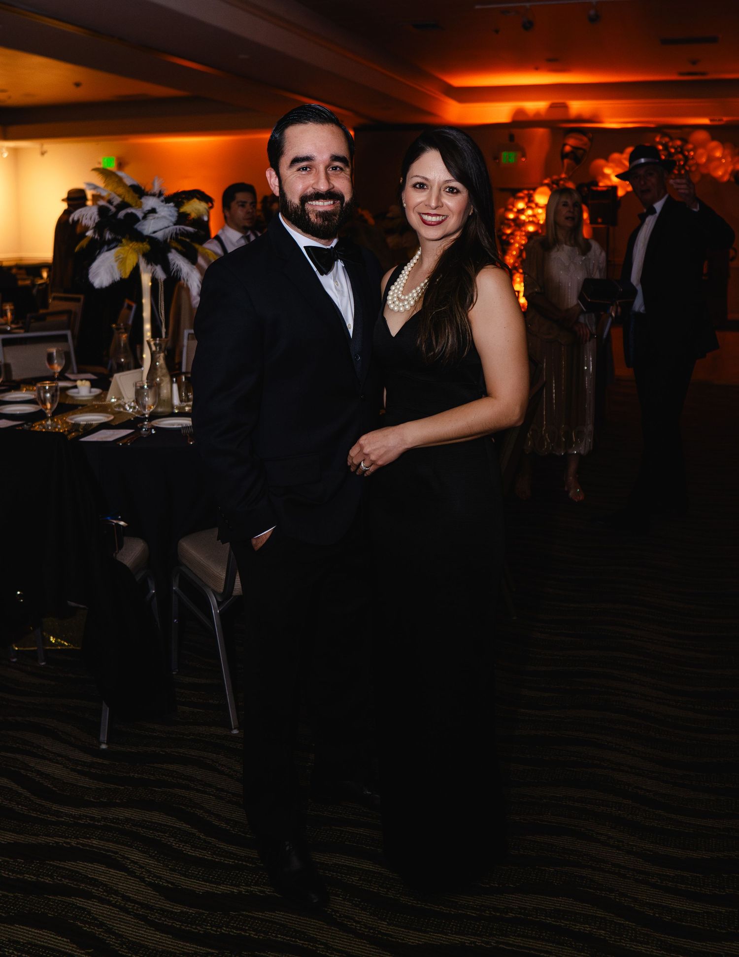 Couple in elegant black formal attire posing together at a glamorous gala event with warm ambient lighting.