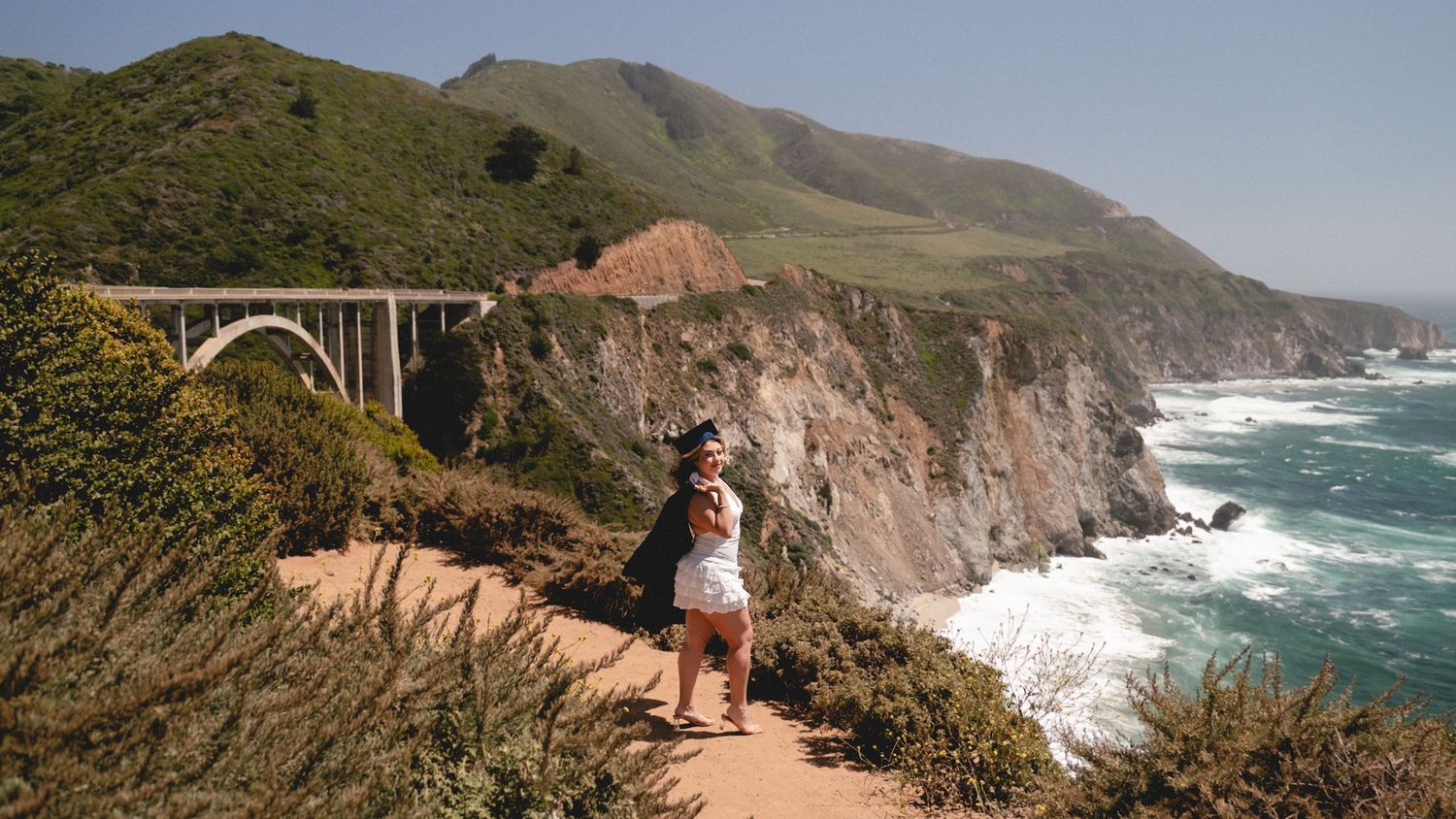 Woman posing on a cliff overlooking Bixby Creek Bridge and the rugged Big Sur coastline in California.