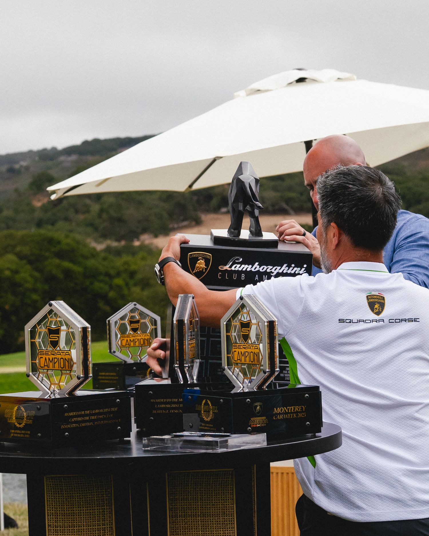 Two men arrange Lamborghini Squadra Corse racing trophies on a table outdoors at an awards event.