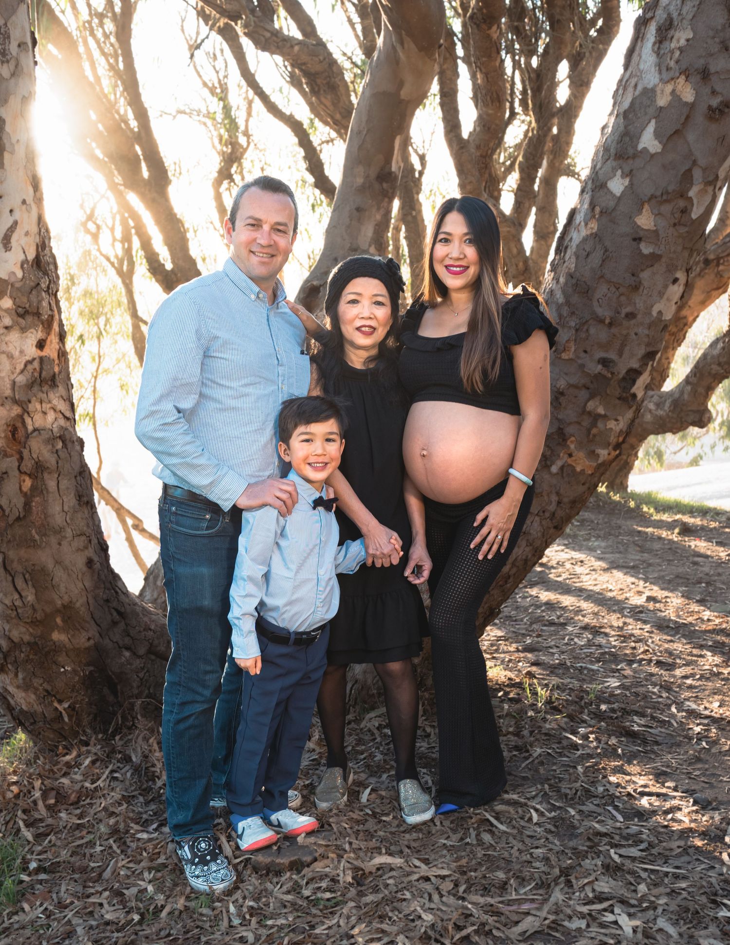 Pregnant woman poses with family including husband and young son in front of large trees during outdoor maternity photo shoot.