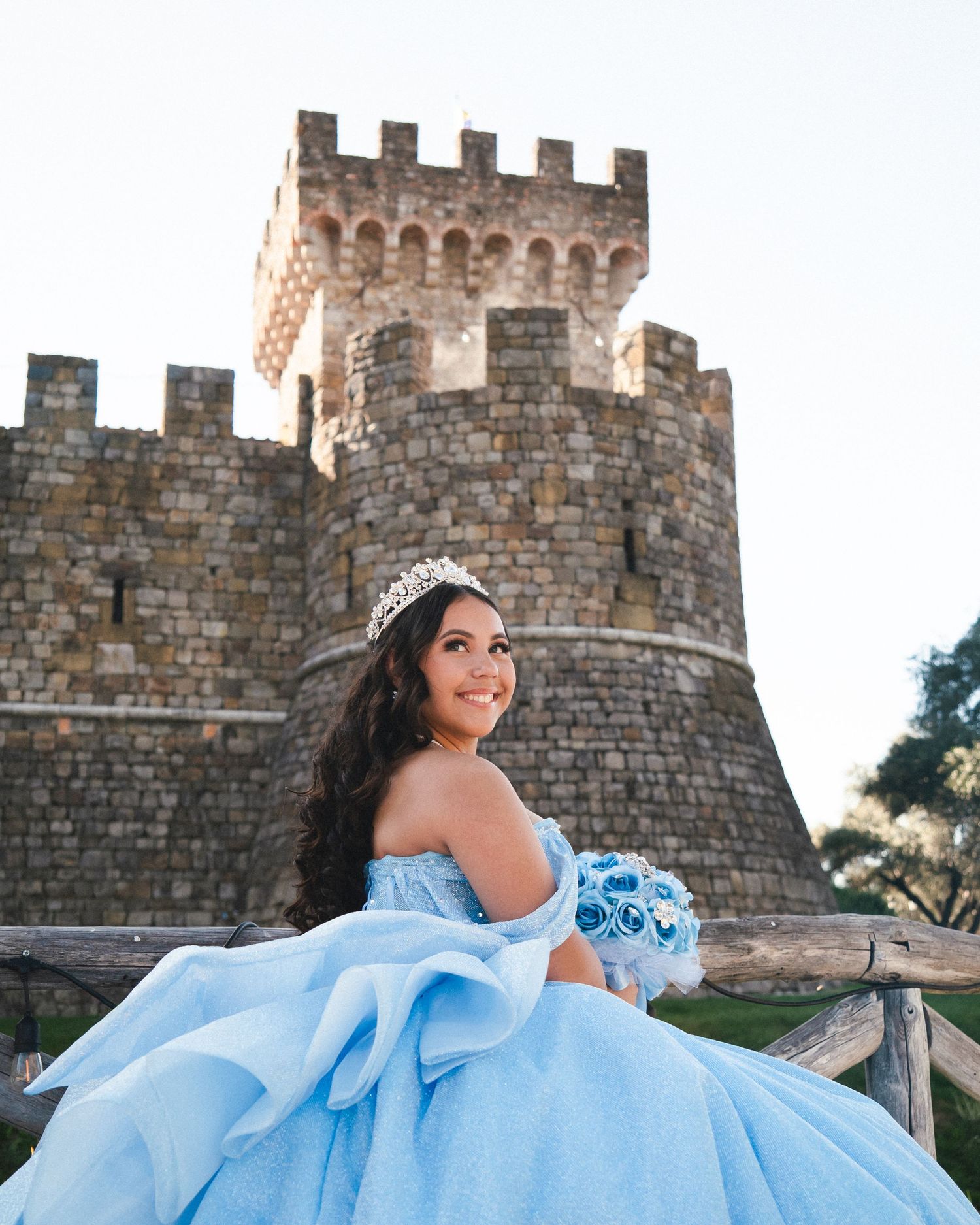 Young woman in blue quinceañera gown and tiara poses with bouquet before a stone castle.