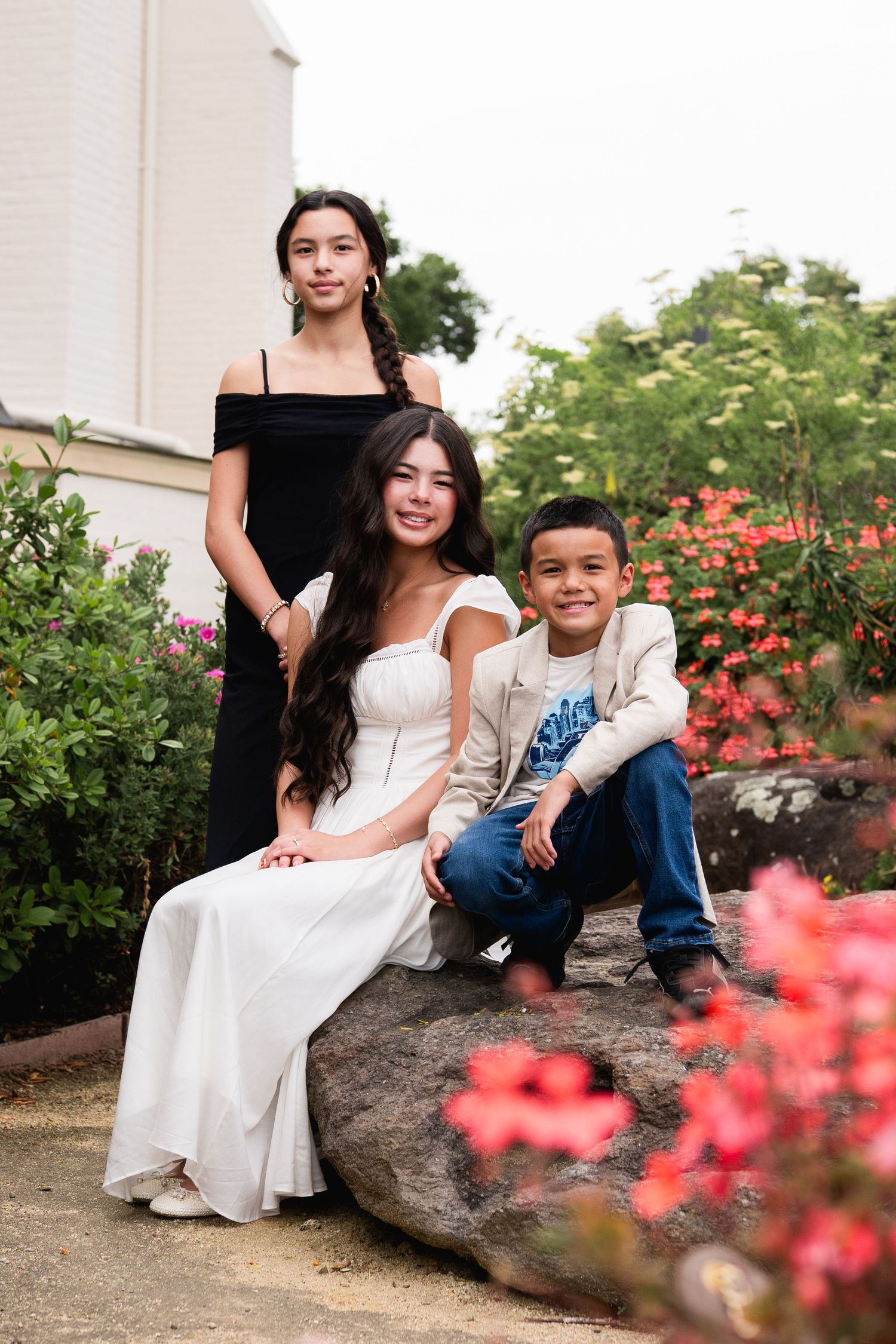 Three siblings in formal wear posing on a rock surrounded by colorful garden flowers.