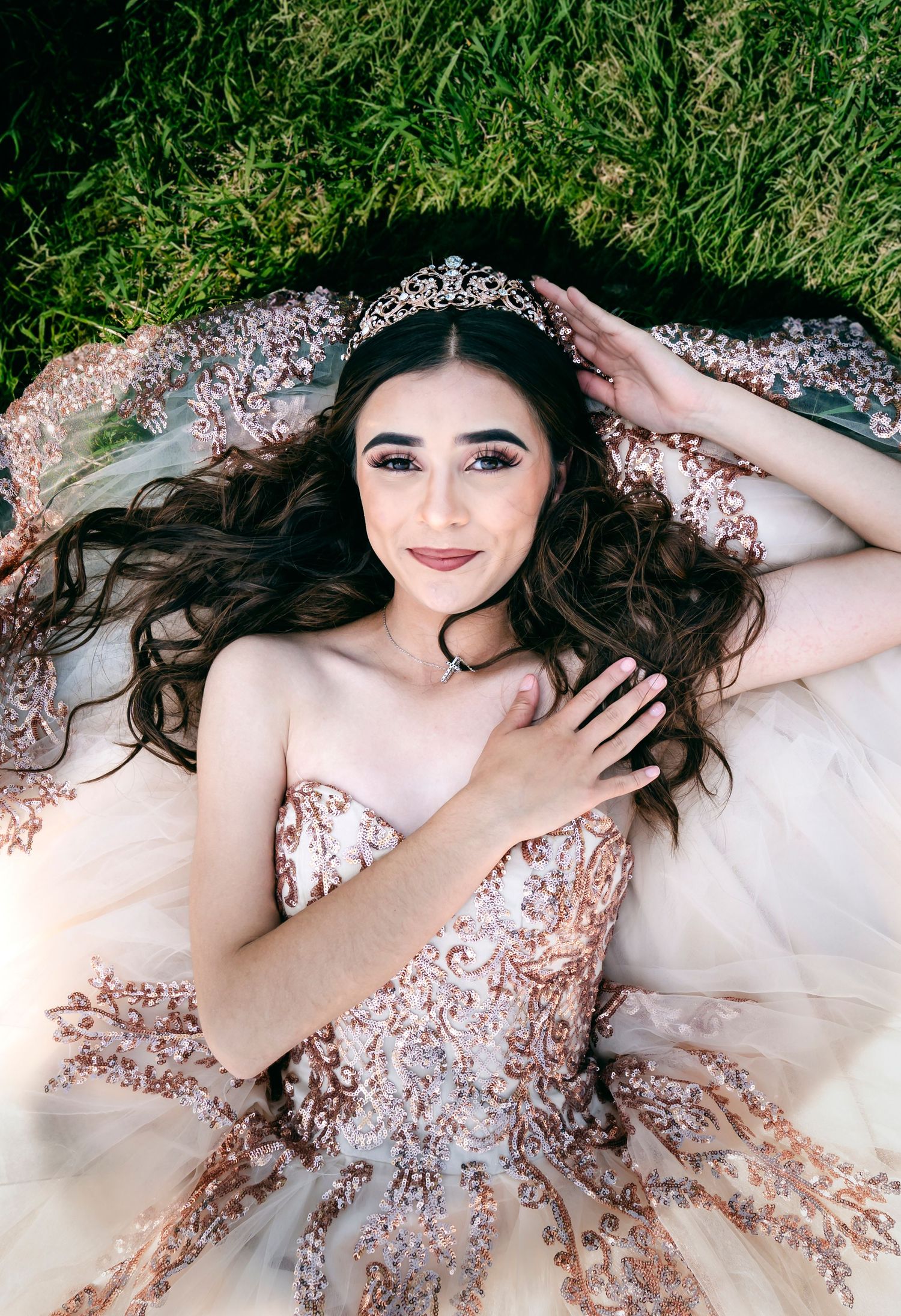 Elegant rose gold sequined gown with tiara sparkles against green grass as long dark hair fans out on white fabric.