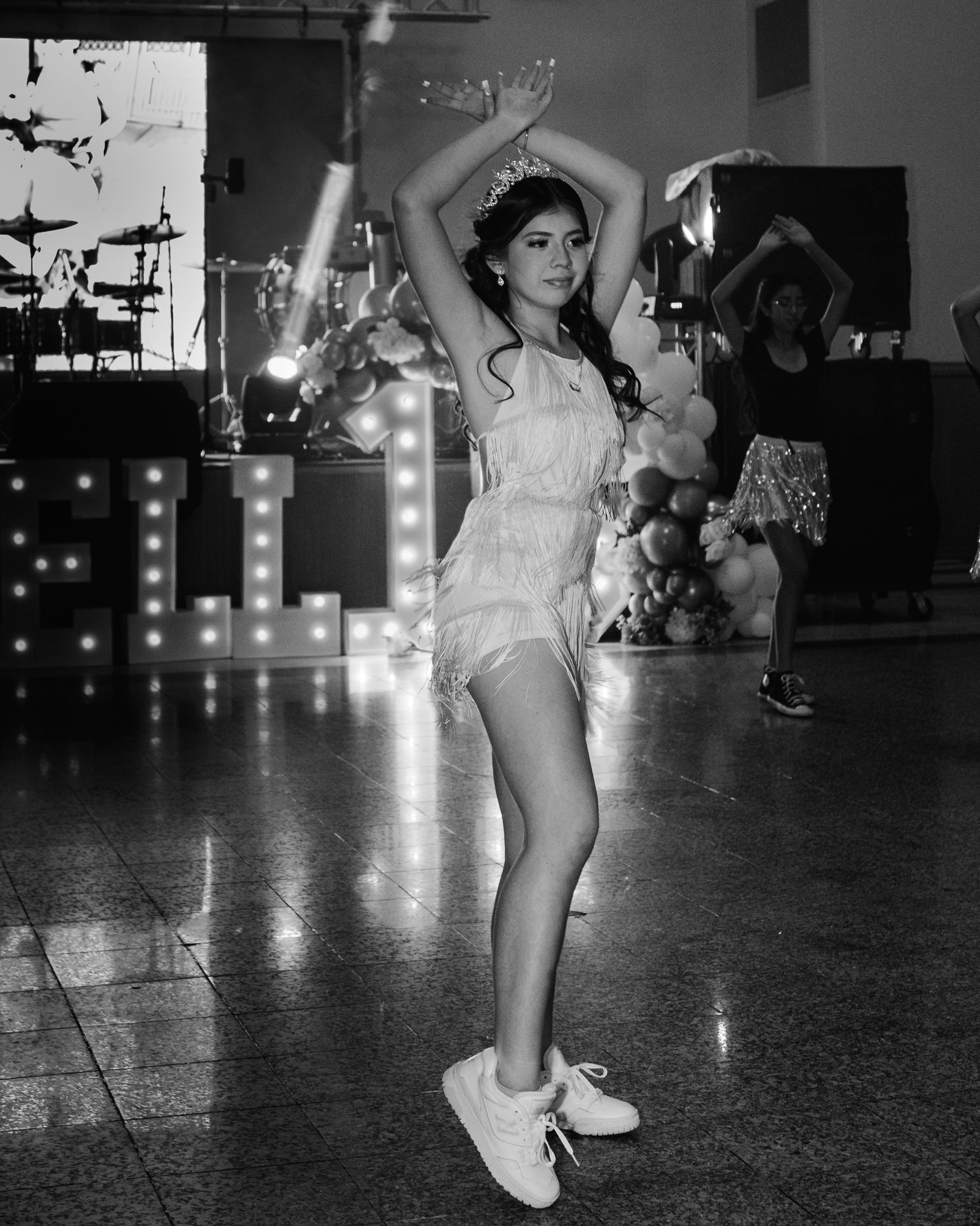 Young woman in white fringe dress and tiara dancing at a festive event with marquee letters in background.