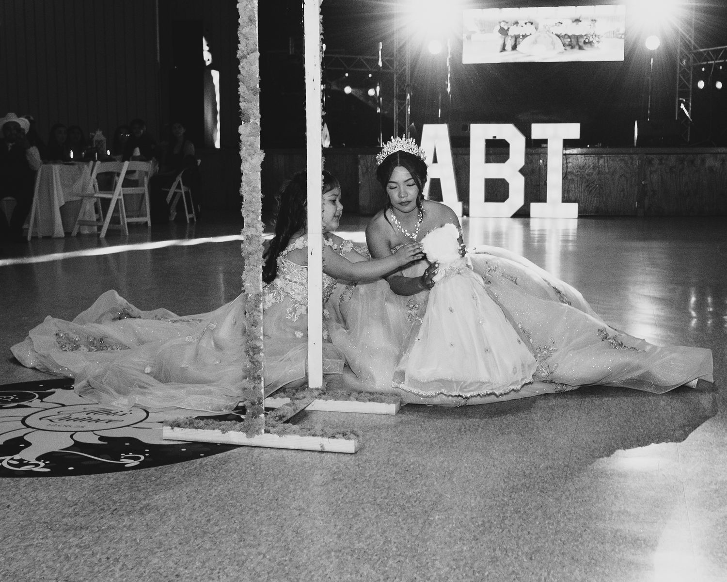 Black and white photo of quinceañera girl in elegant gown posing near illuminated letter backdrop on dance floor.