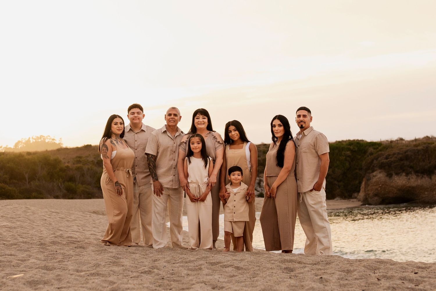 A group of people in neutral-toned attire pose together on a scenic beach at sunset.