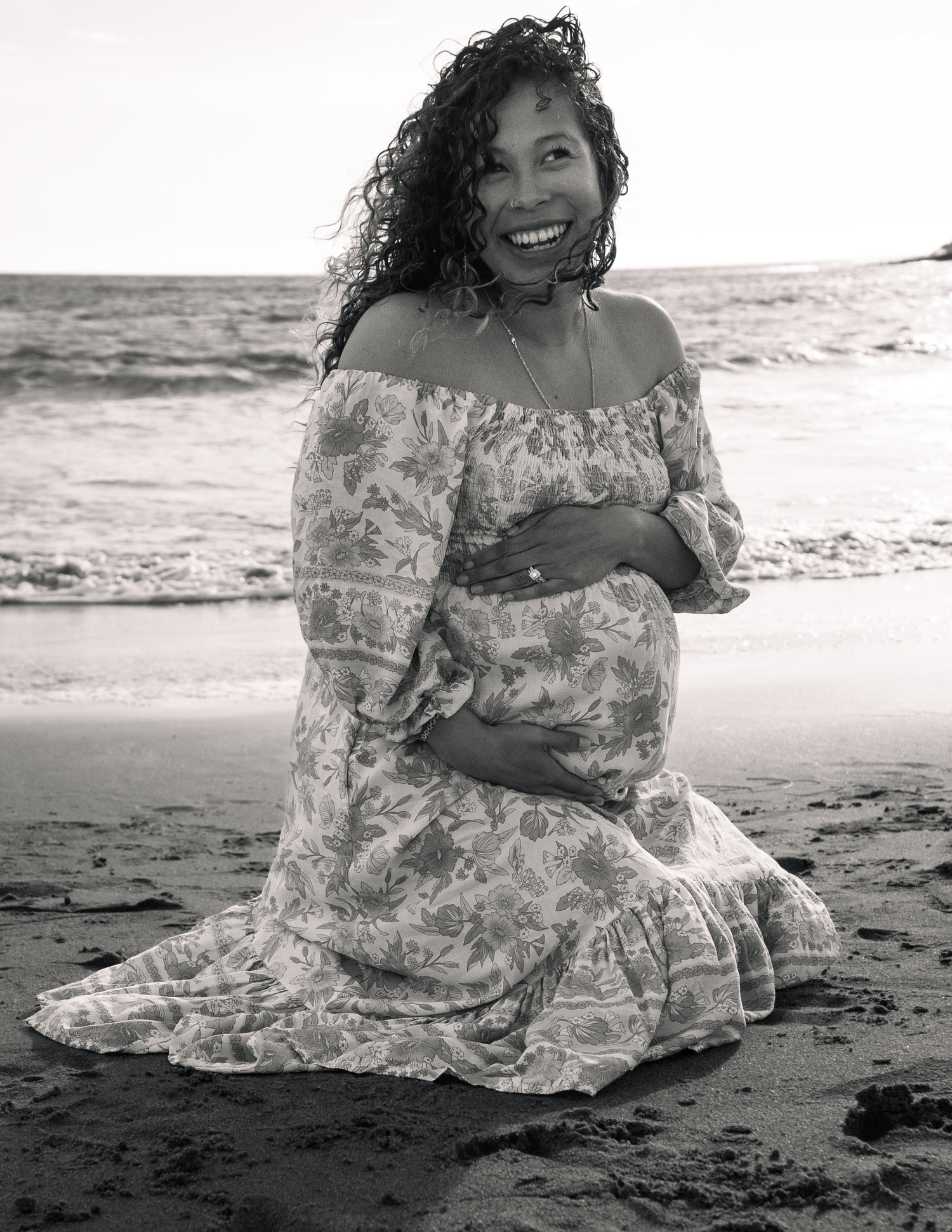 Black and white photo of a smiling pregnant woman in a floral dress kneeling on a beach, cradling her baby bump.