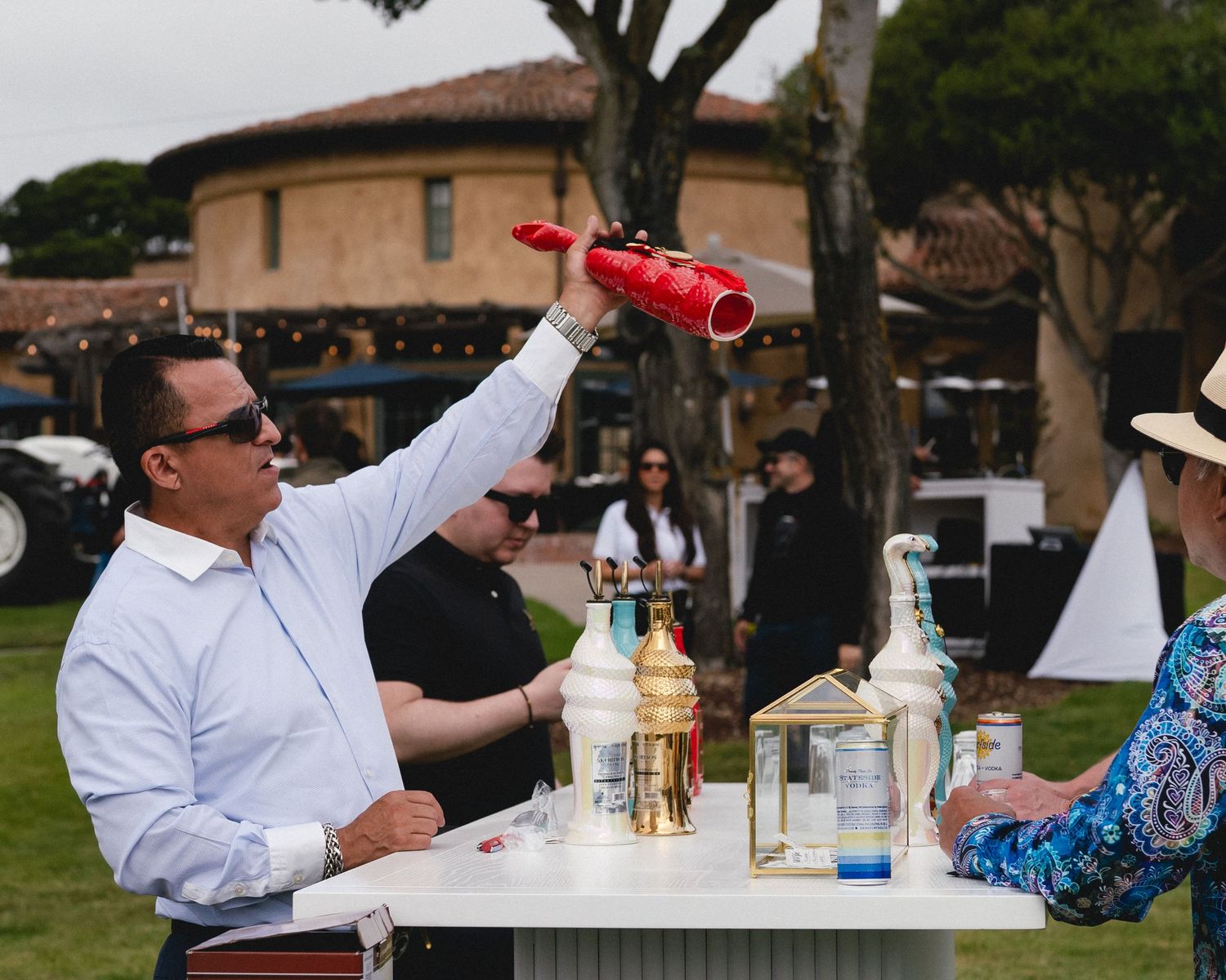 Bartender pouring cocktail from red shaker at outdoor bar event on green lawn.