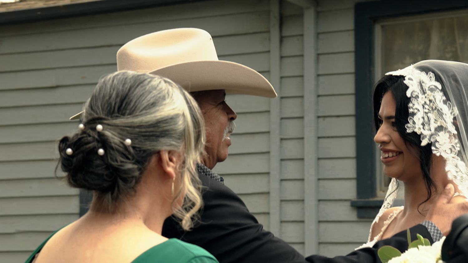 A bride in a white veil holds flowers while greeting guests at an outdoor wedding, with a man in a cowboy hat nearby.
