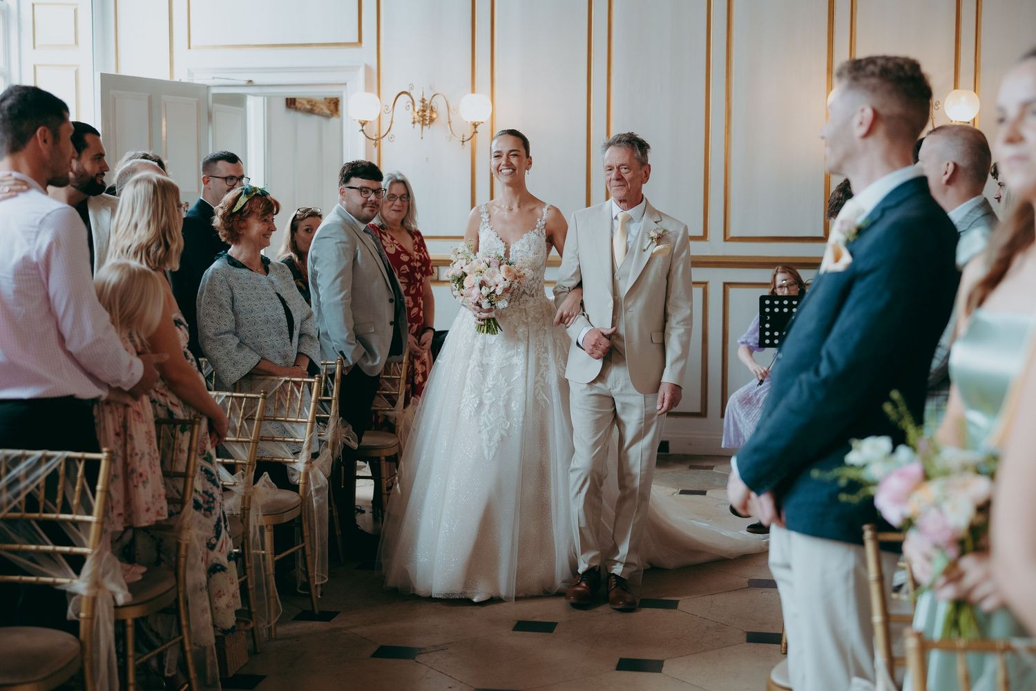 Bride walks down the aisle in a white gown with bouquet, escorted by a man in a beige suit at an elegant wedding ceremony.