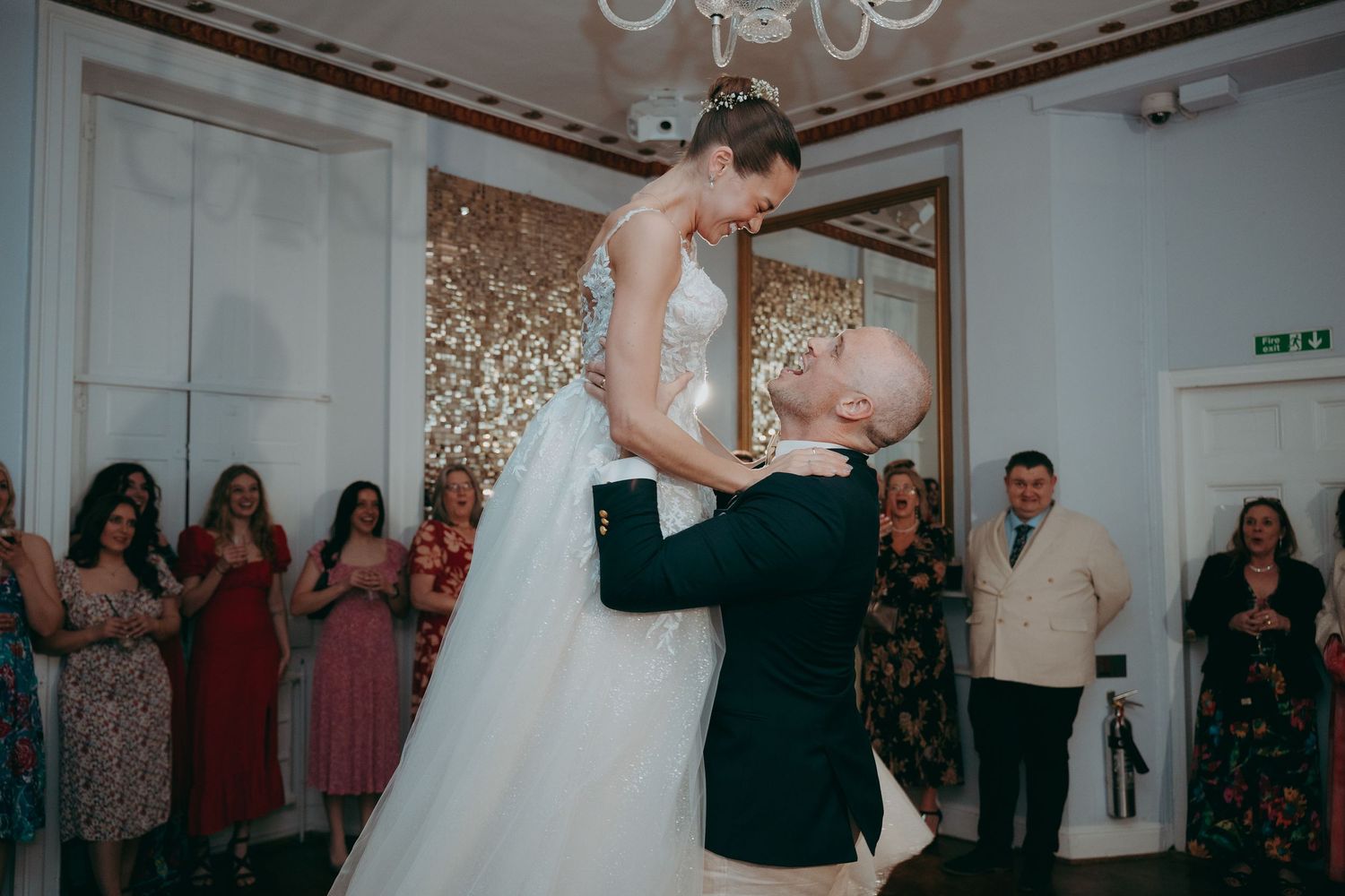 Groom lifts bride in white wedding dress during first dance at elegant wedding reception with guests applauding.