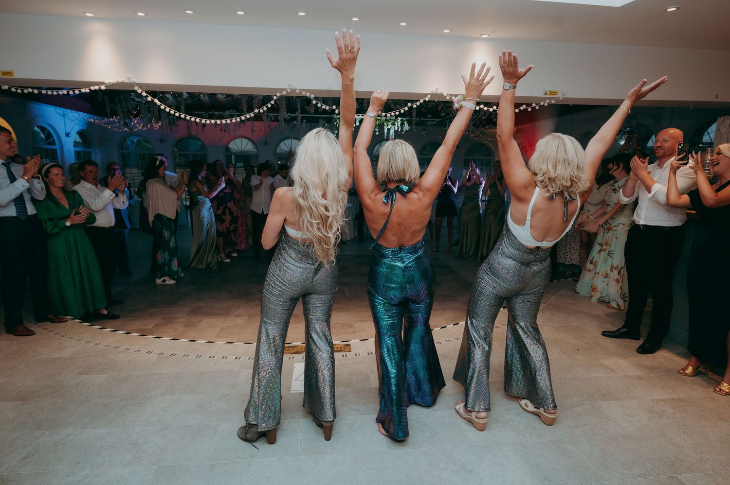Three women in metallic flared jumpsuits dancing with arms raised at a lively wedding reception.