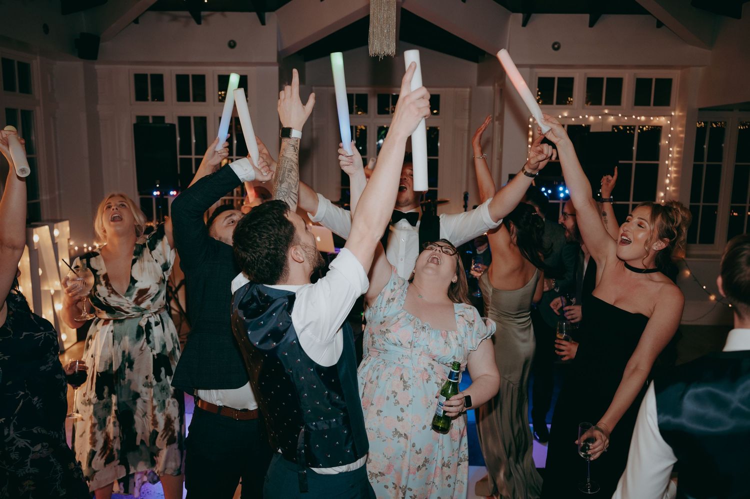 Wedding guests dancing and raising glow sticks at a festive indoor reception celebration.