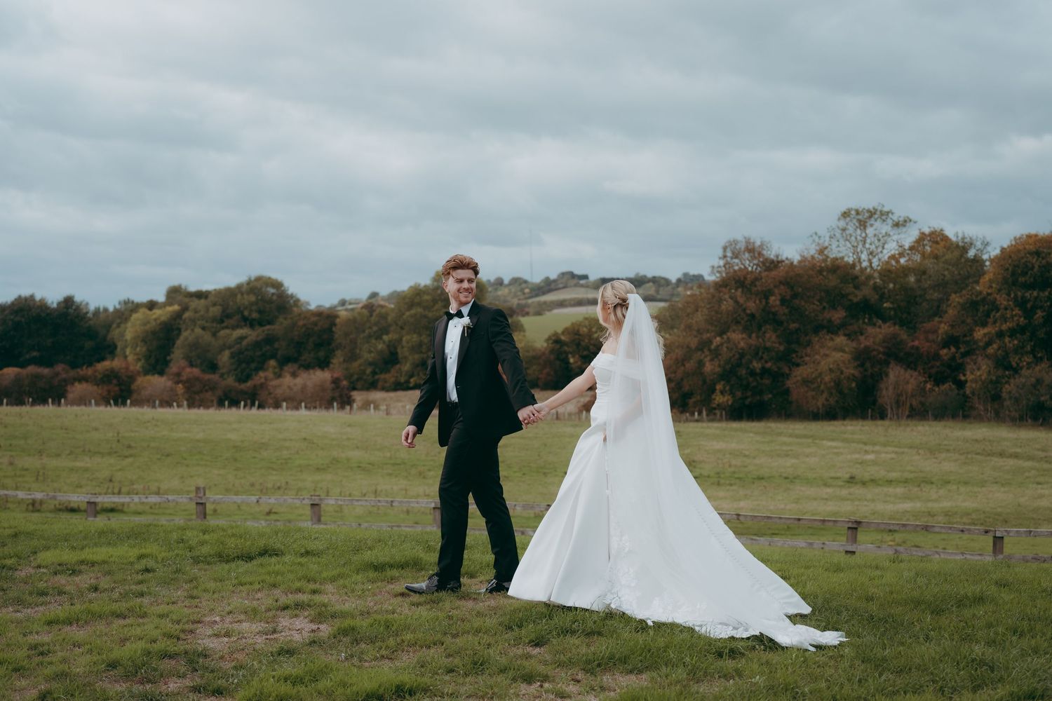 Bride and groom holding hands in a green countryside field, bride wearing a white gown with veil, groom in black tuxedo.
