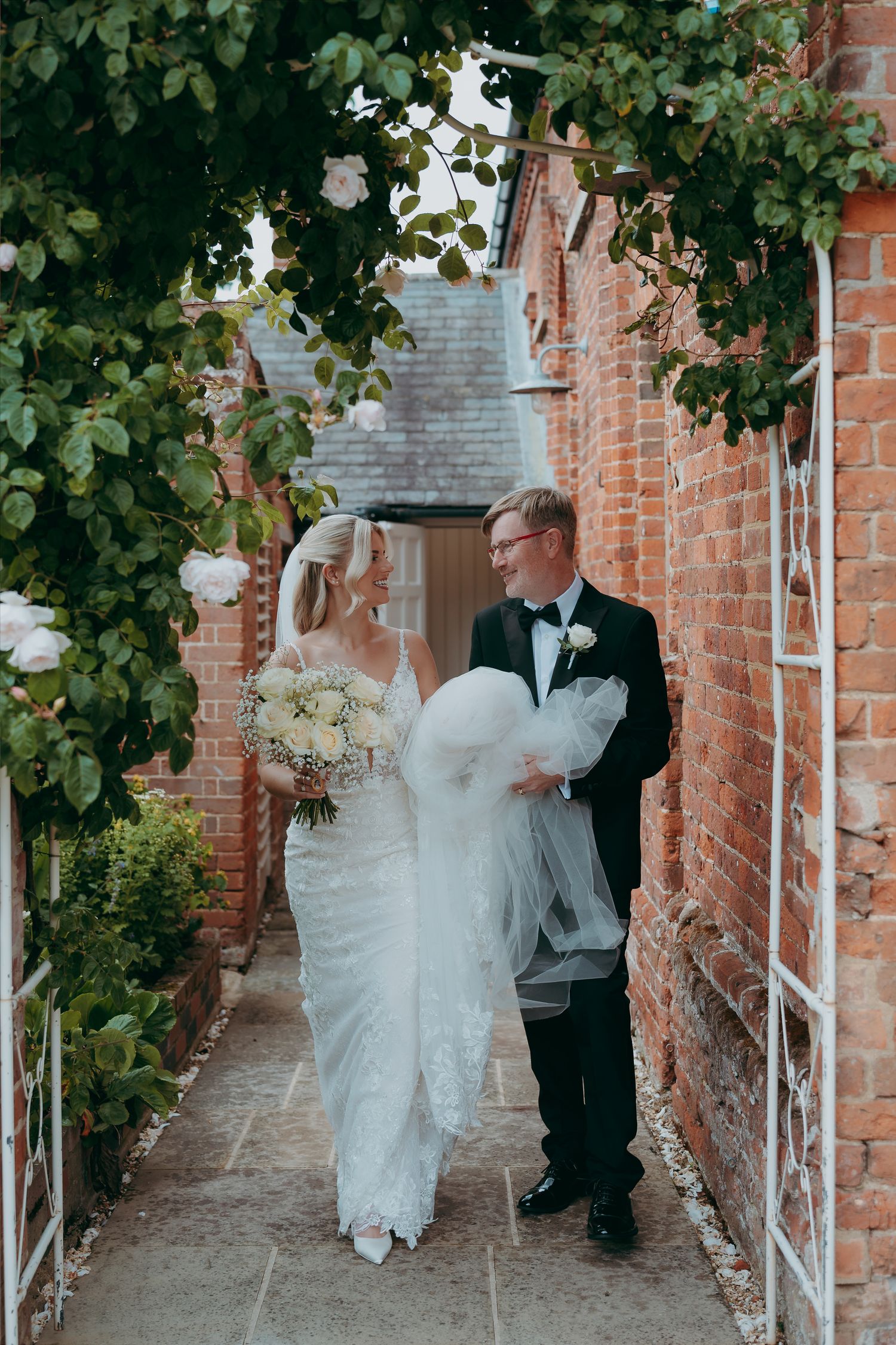 Bride and groom exchange vows at rustic stone wall venue with circular greenery arch and officiant behind them.
