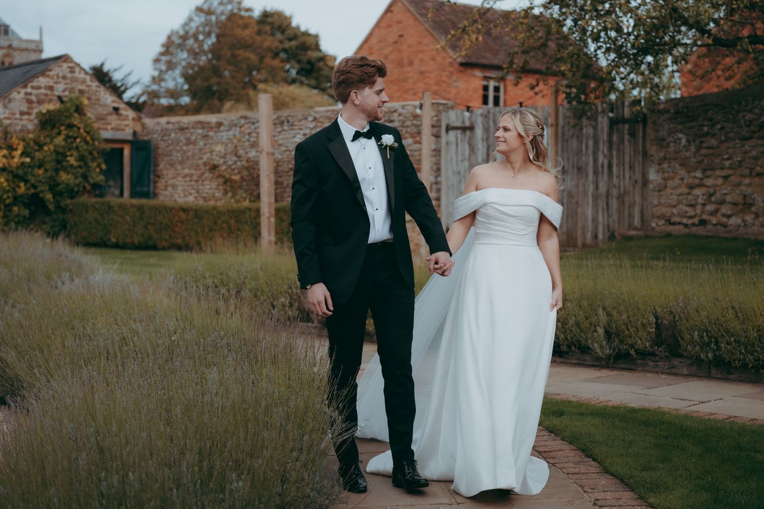 Bride and groom walk hand in hand through a garden path at a rustic countryside wedding venue.