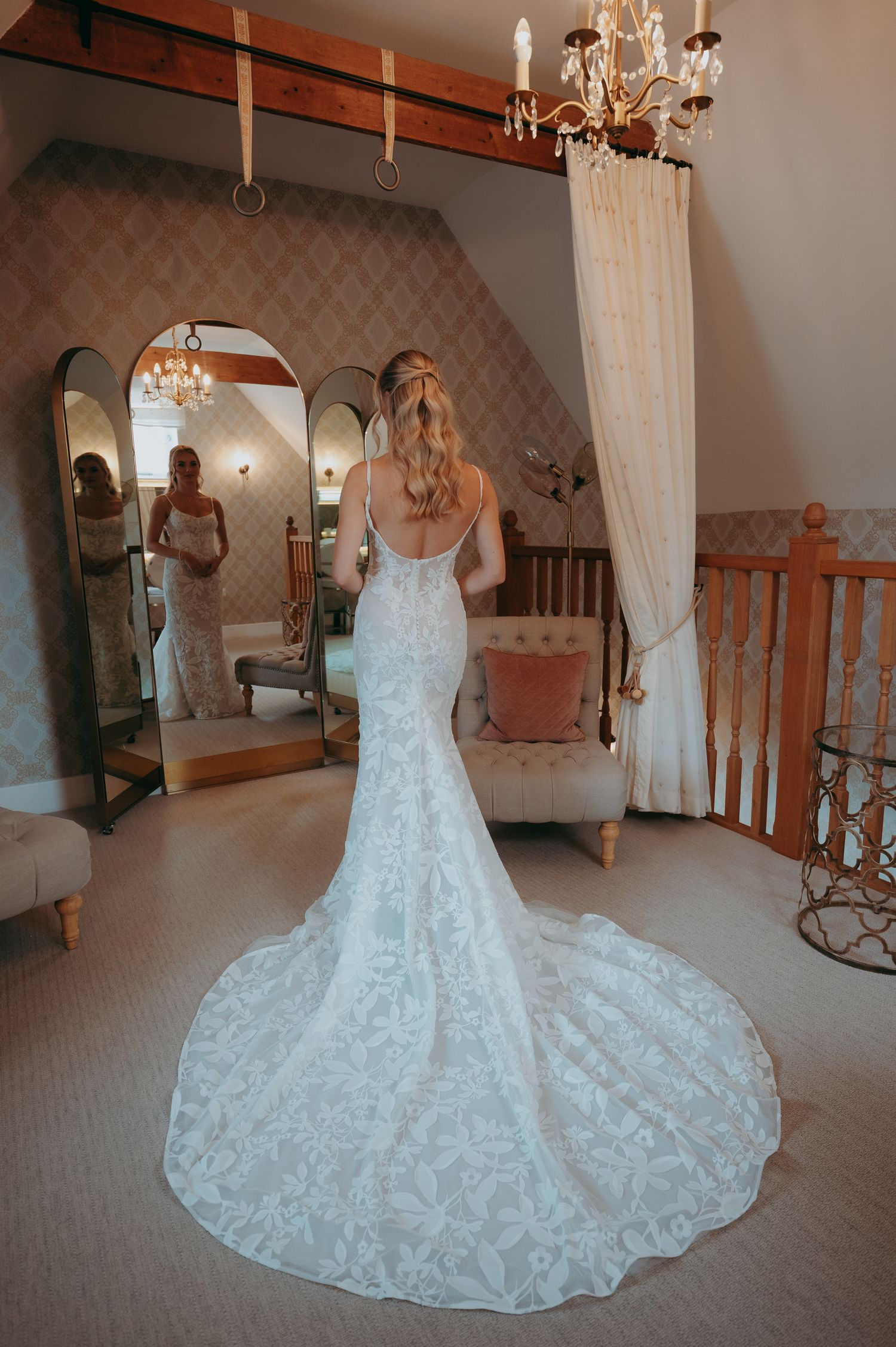 Bride in elegant lace mermaid wedding gown with long train, viewed from behind in a charming bridal suite.