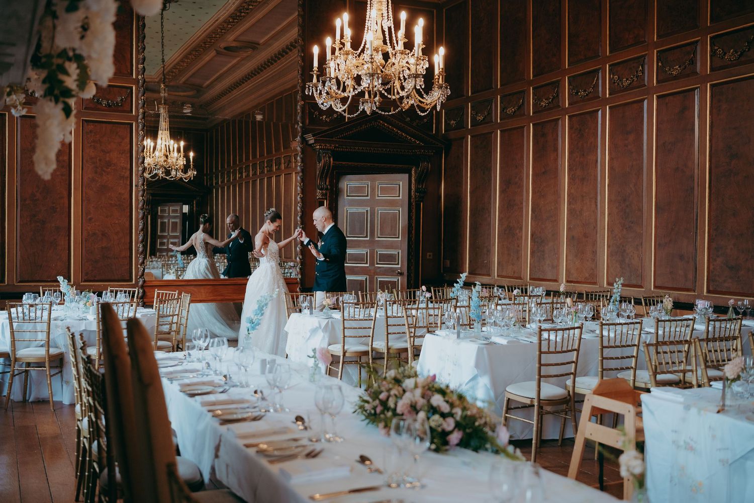 Elegant wedding reception in ornate wood-paneled ballroom with chandeliers, chiavari chairs, and couple sharing first dance.