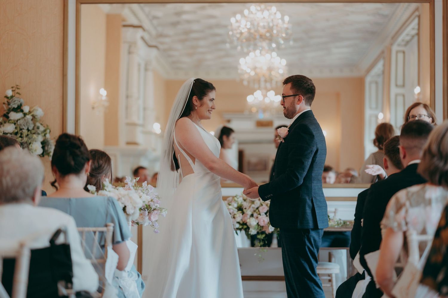 Bride and groom holding hands during elegant wedding ceremony in a grand ballroom with chandelier.