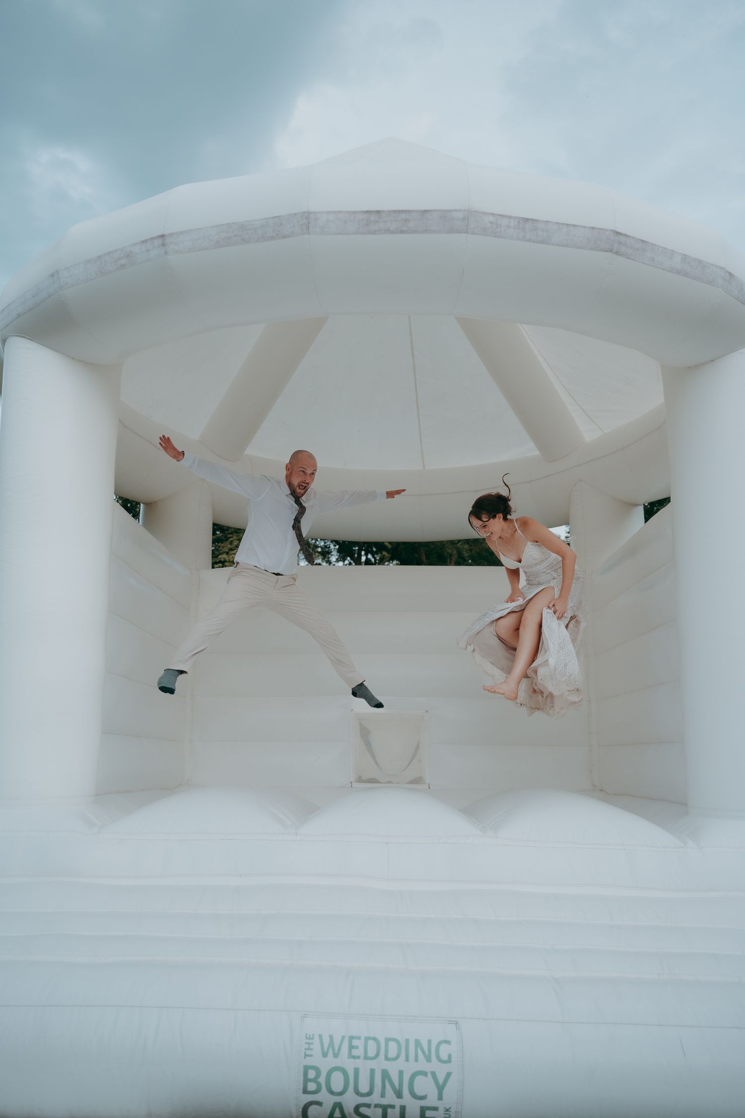 Bride and groom jumping joyfully inside a white Wedding Bouncy Castle, wearing formal attire under a cloudy sky.