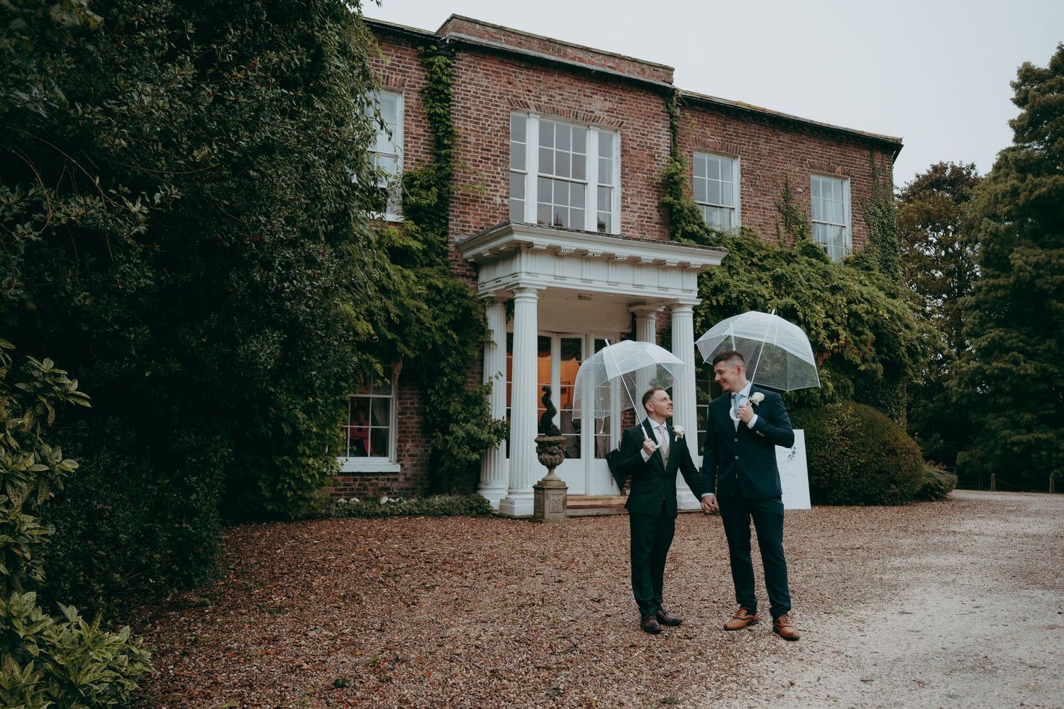 Two grooms in suits holding clear umbrellas outside a grand brick manor house on a rainy wedding day.
