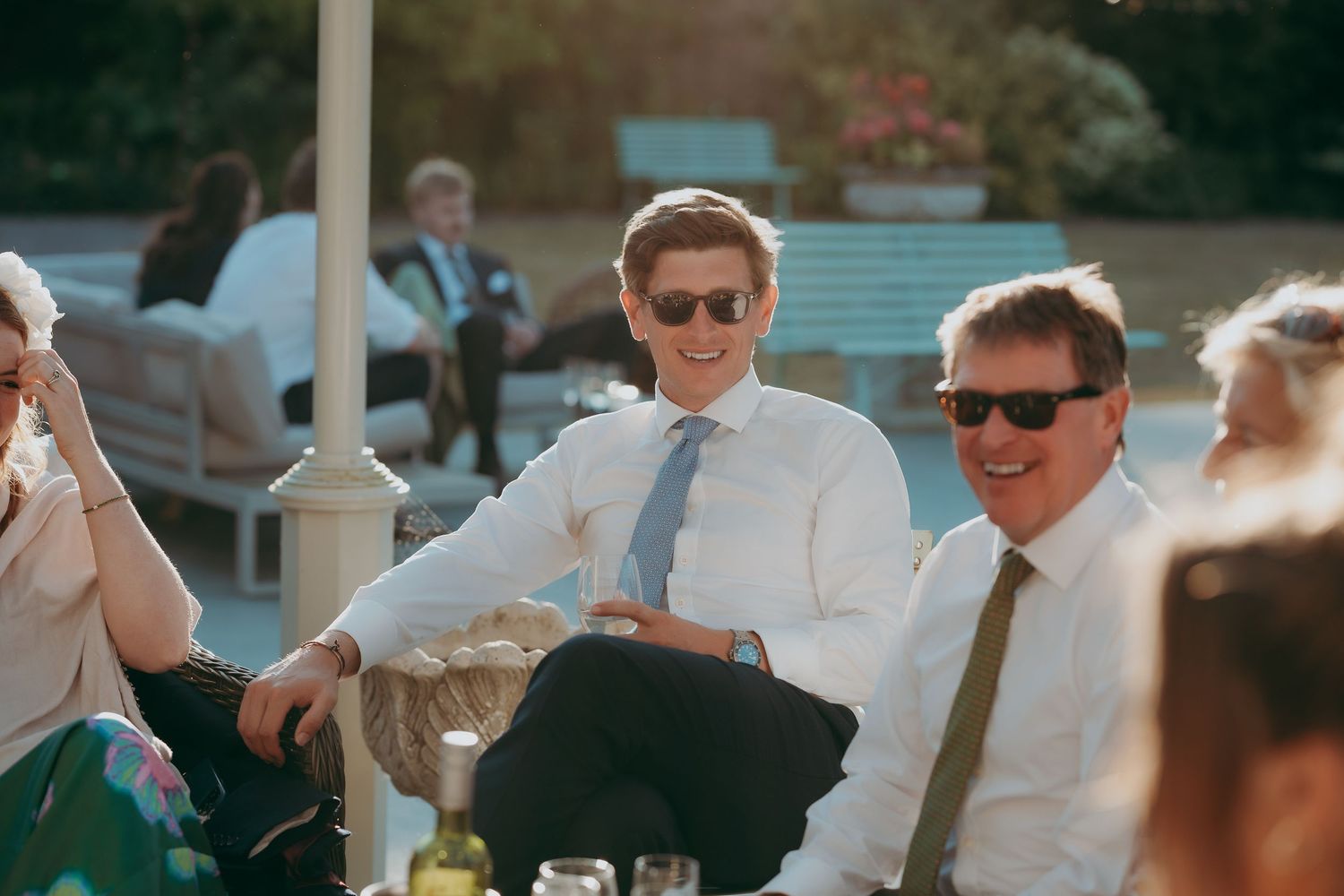Two men in suits and sunglasses smiling at an outdoor wedding reception on a sunny day.
