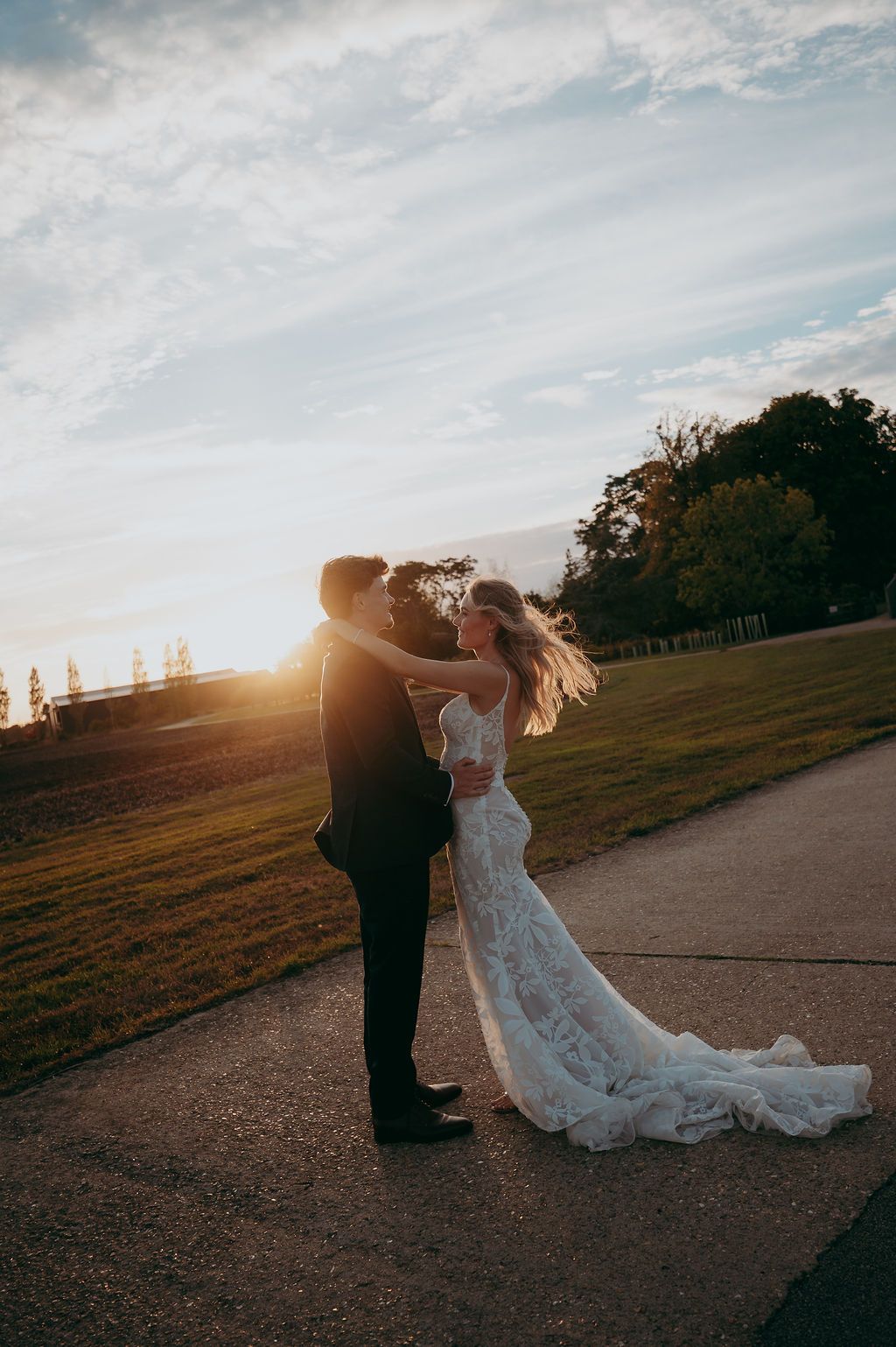 Bride in a lace wedding gown embraces her groom in a dark suit on a paved path at golden hour sunset.