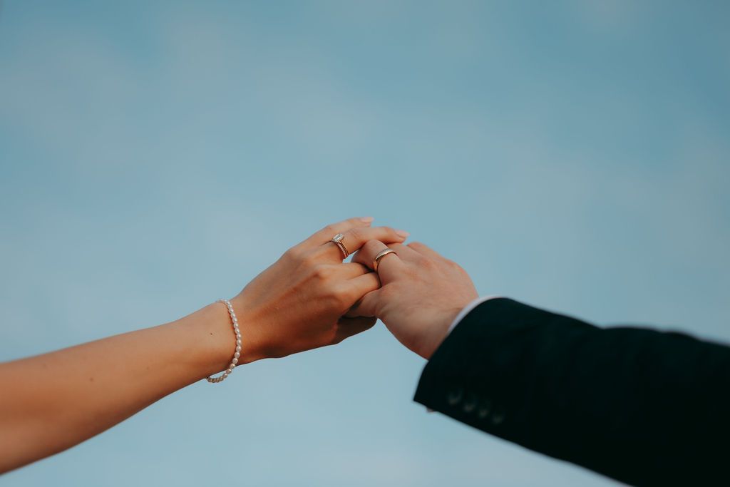 Bride and groom holding hands with wedding rings visible against a soft blue sky, her bracelet and his dark suit sleeve in view.