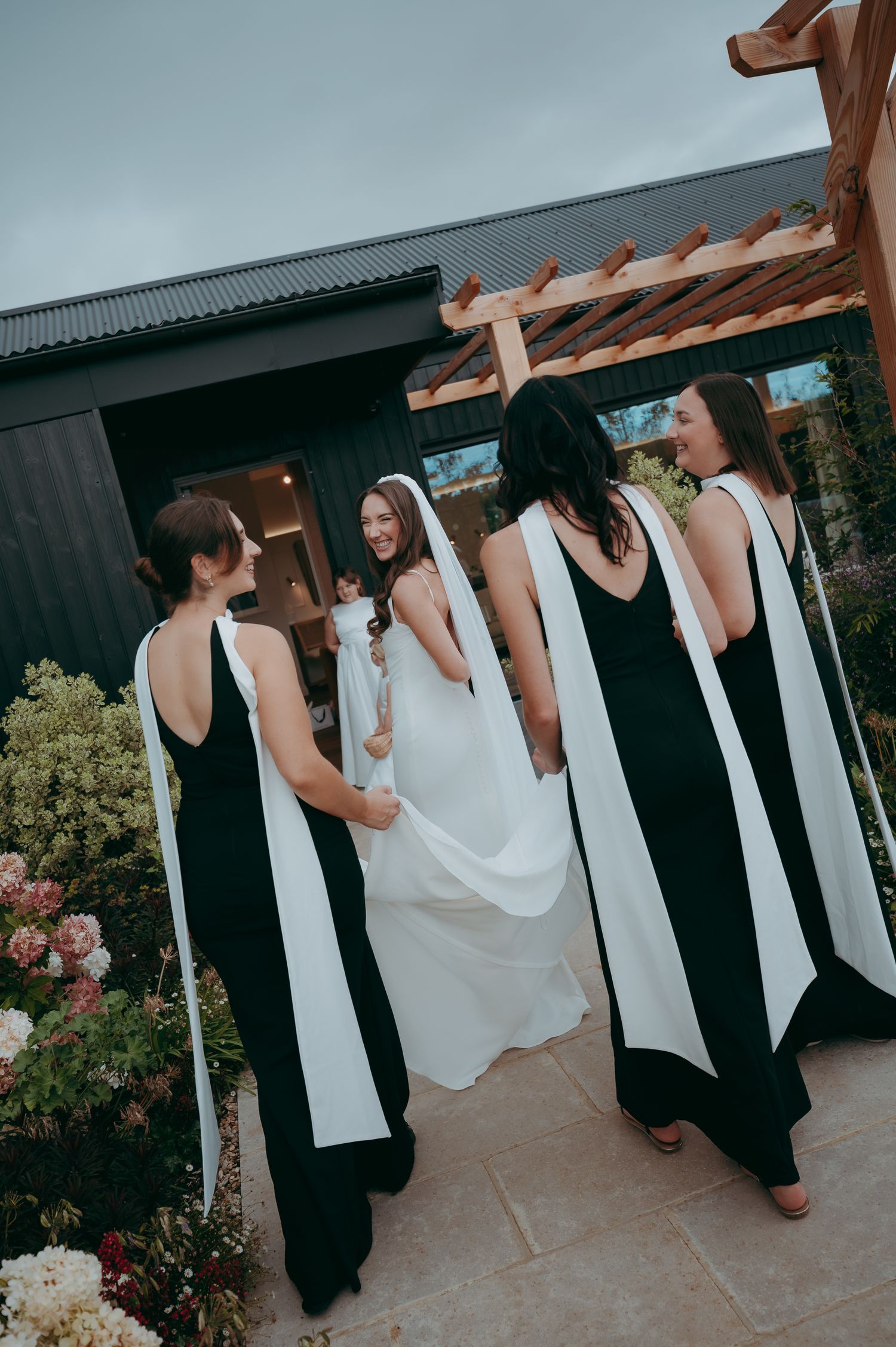 Bride in white gown with bridesmaids in black dresses with white sashes outside a modern venue.