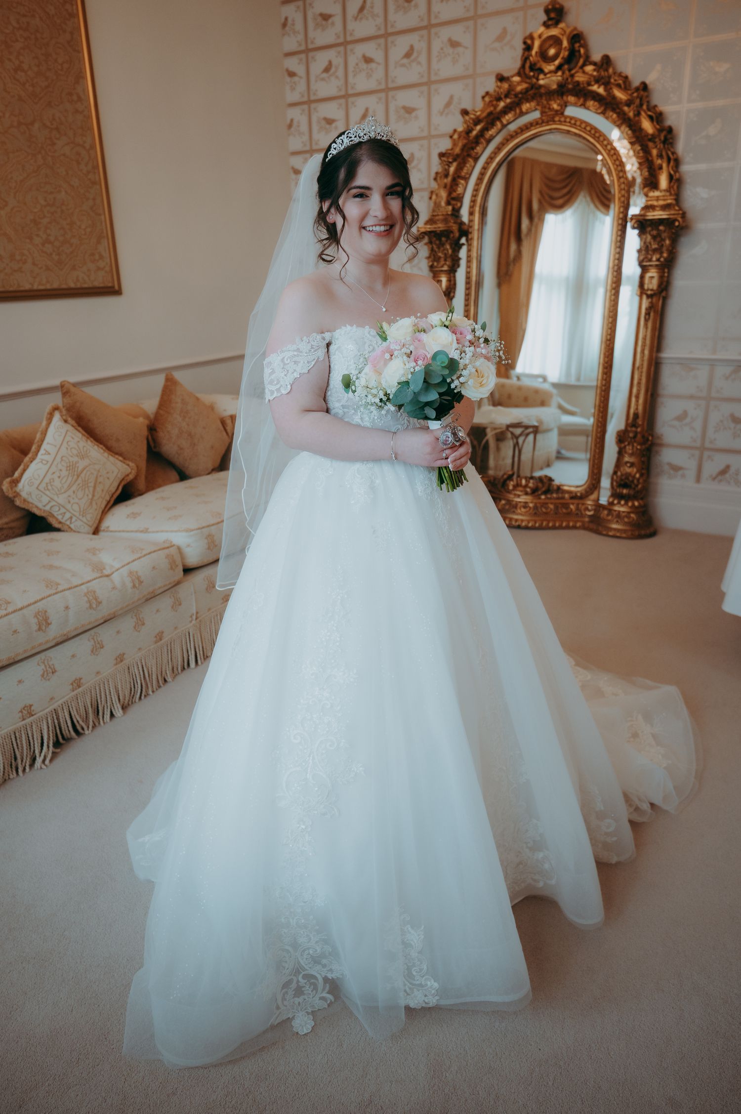 Bride in off-shoulder white ballgown with veil and tiara holds bouquet before ornate gold mirror in elegant room.