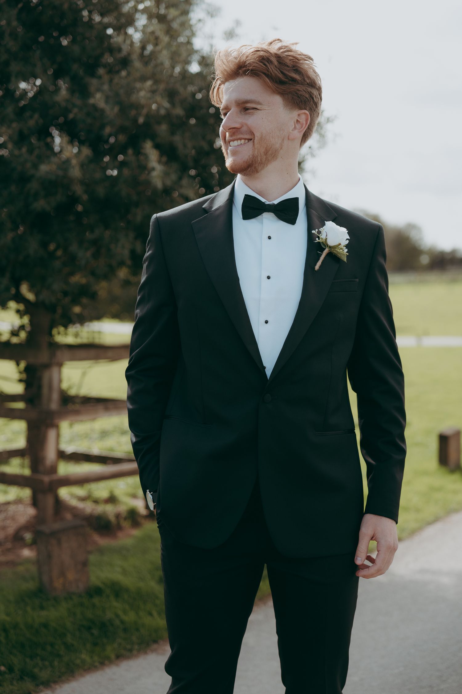 Smiling groom in black tuxedo with bow tie and white boutonniere standing outdoors on a path at a wedding venue.