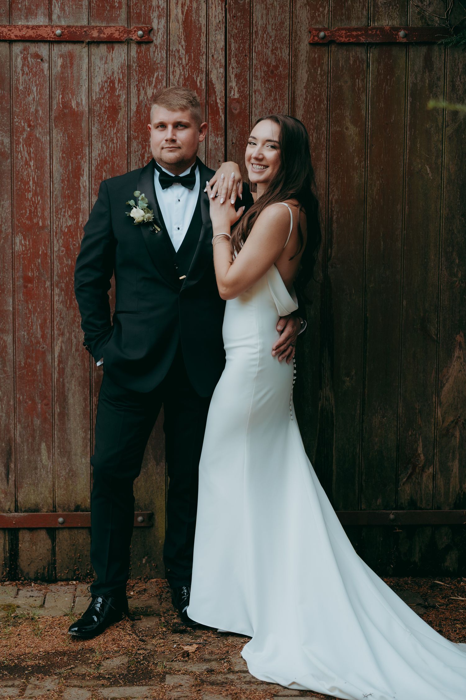 Bride in white gown and groom in black tuxedo posing together in front of rustic red barn doors.