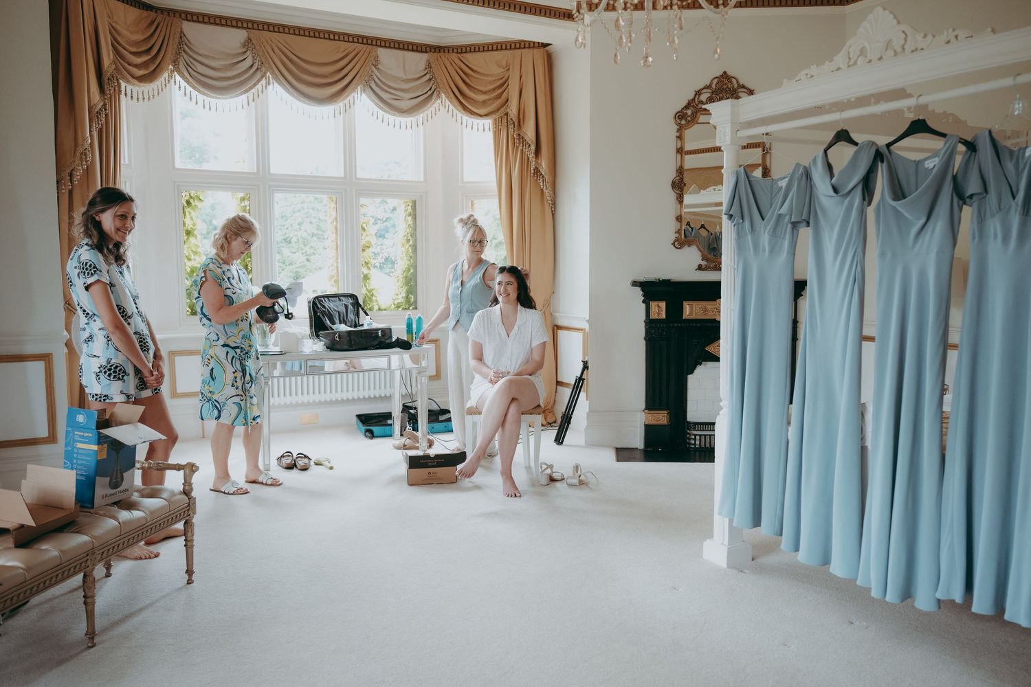 Bridesmaids preparing in elegant room with light blue dresses hanging, photographer capturing candid getting-ready moments.
