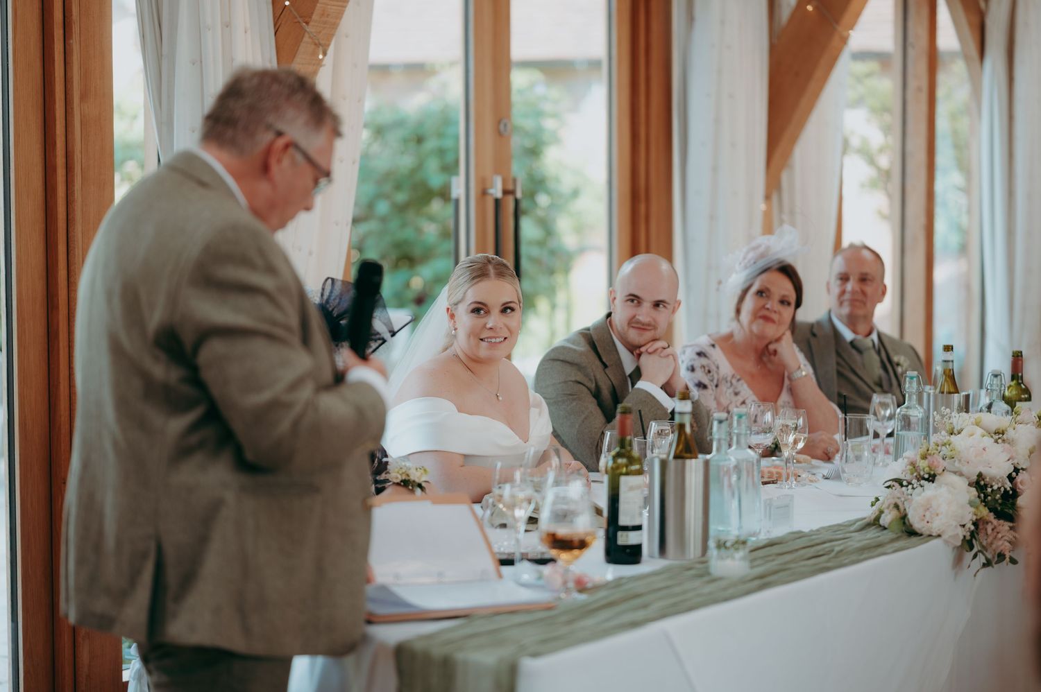 Wedding party of five posing before rustic brick wall, bride in white gown with bouquet, groom in grey suit, guests in sage green.