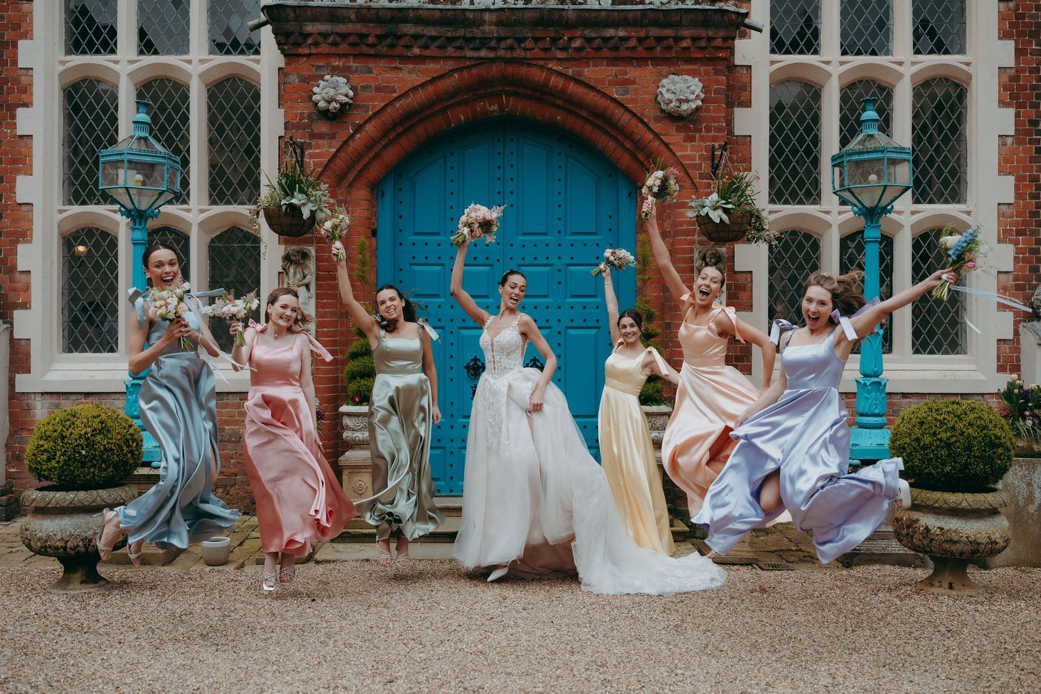 Bride and bridesmaids in colorful satin dresses jumping joyfully outside a historic manor with a blue door.
