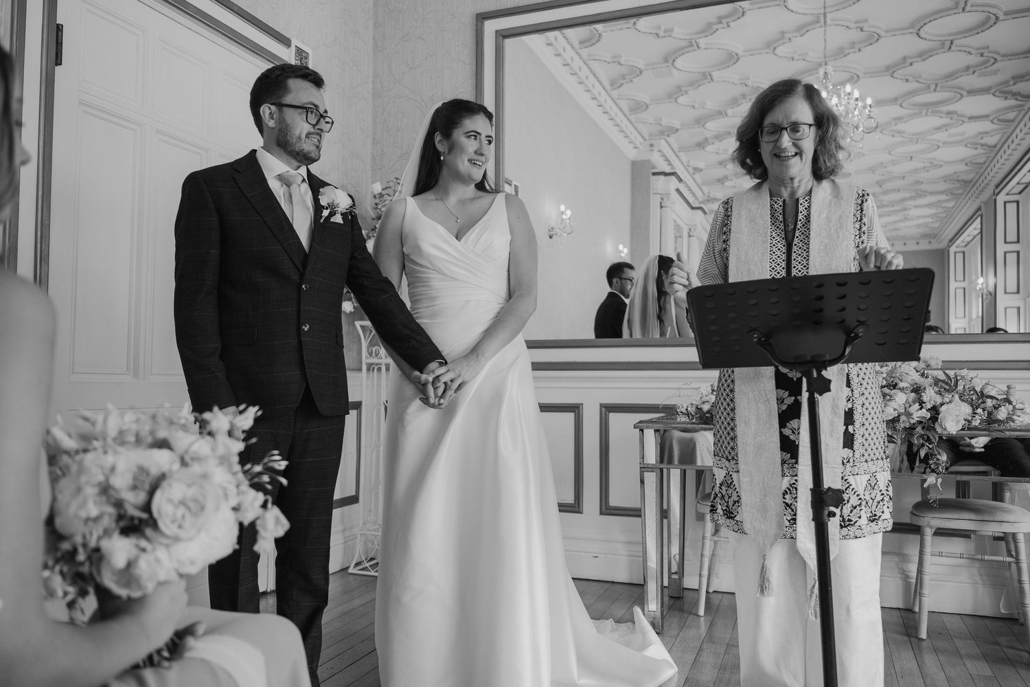 Black and white wedding ceremony photo of smiling bride and groom standing before officiant at elegant venue.