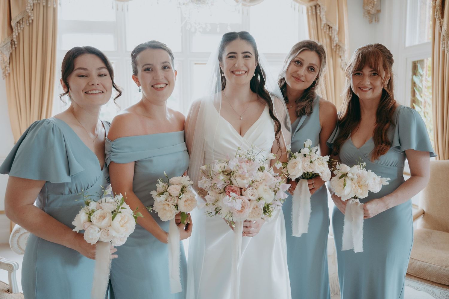 Bride in white gown with four bridesmaids in dusty blue dresses holding white floral bouquets indoors.