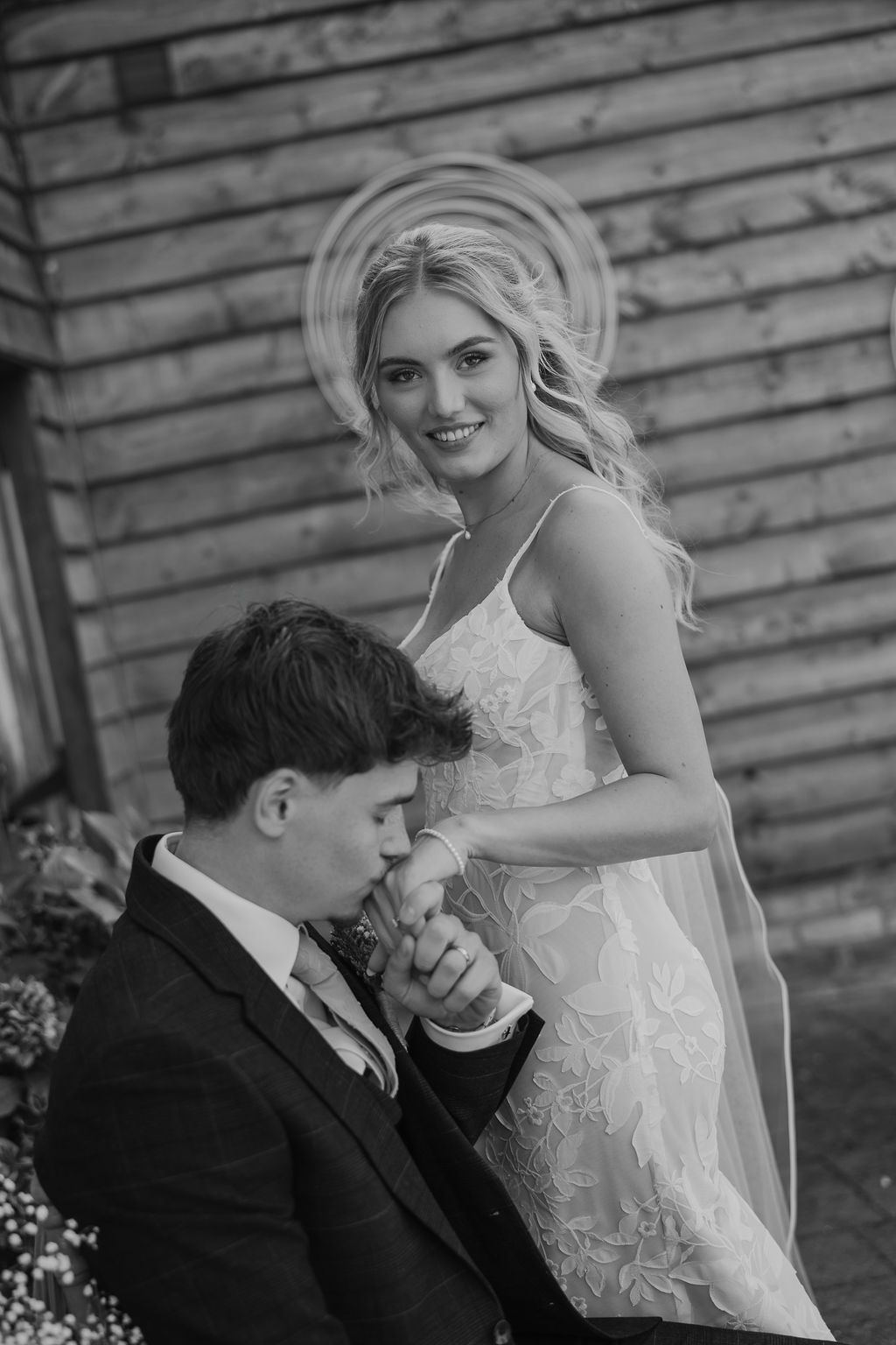 Black and white photo of a smiling bride in a lace wedding gown with a veil as the groom in a suit kisses her hand beside a wooden wall.