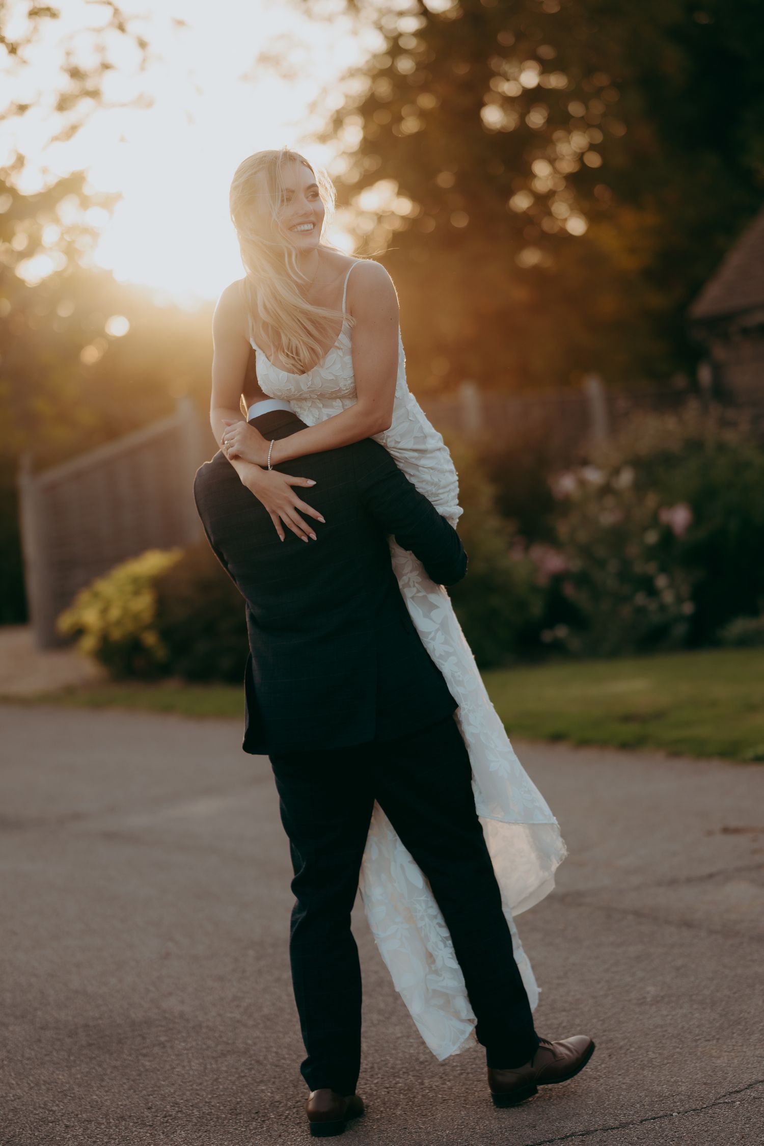 Groom lifting bride in white lace dress during golden hour sunset outdoors.