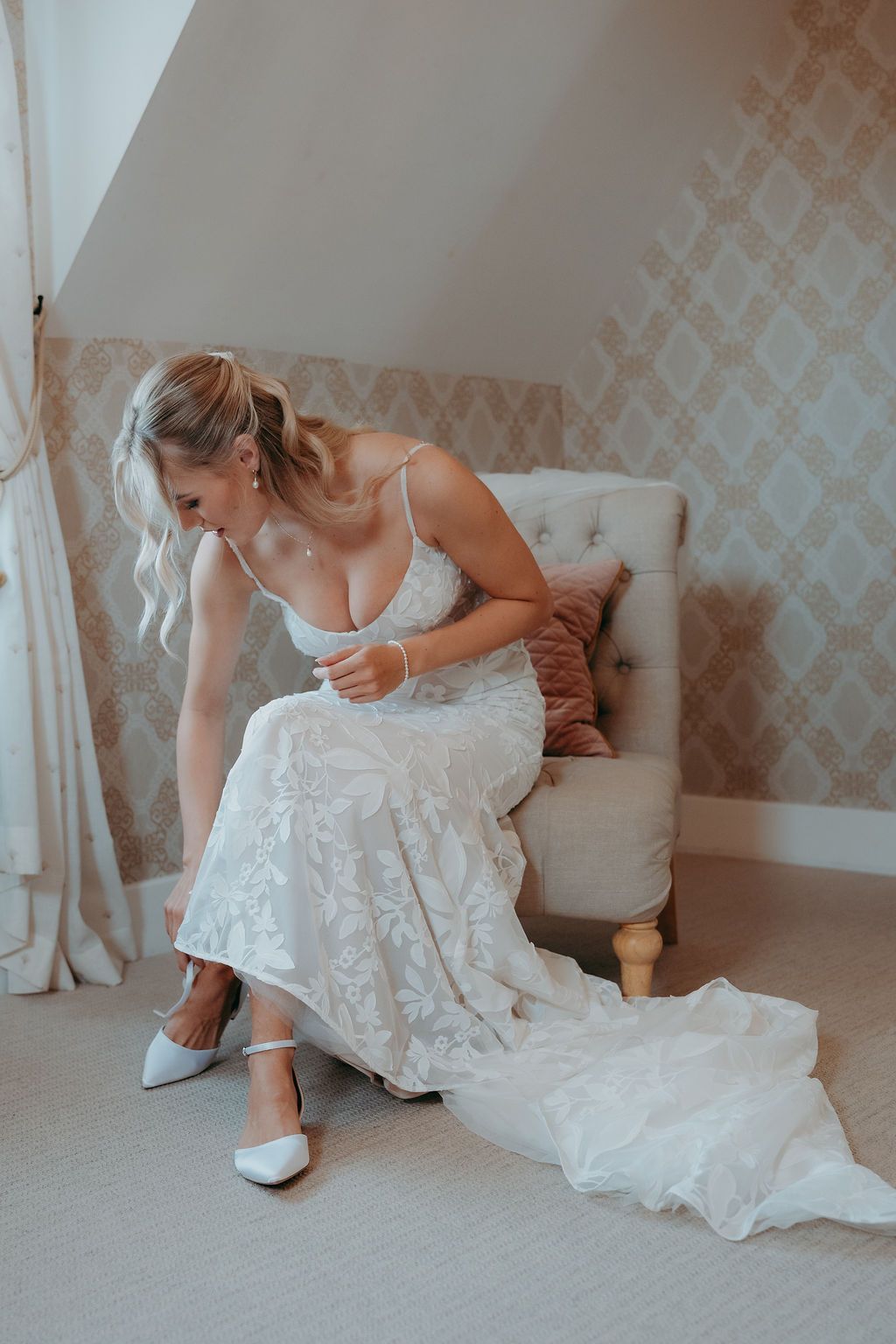 Bride in a lace wedding gown sits on a cushioned chair adjusting her white flat shoe in a softly lit bedroom with patterned wallpaper.