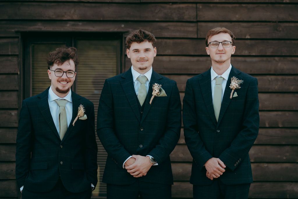 Three groomsmen in navy suits with sage green ties and floral boutonnieres pose side by side in front of a rustic wooden wall.