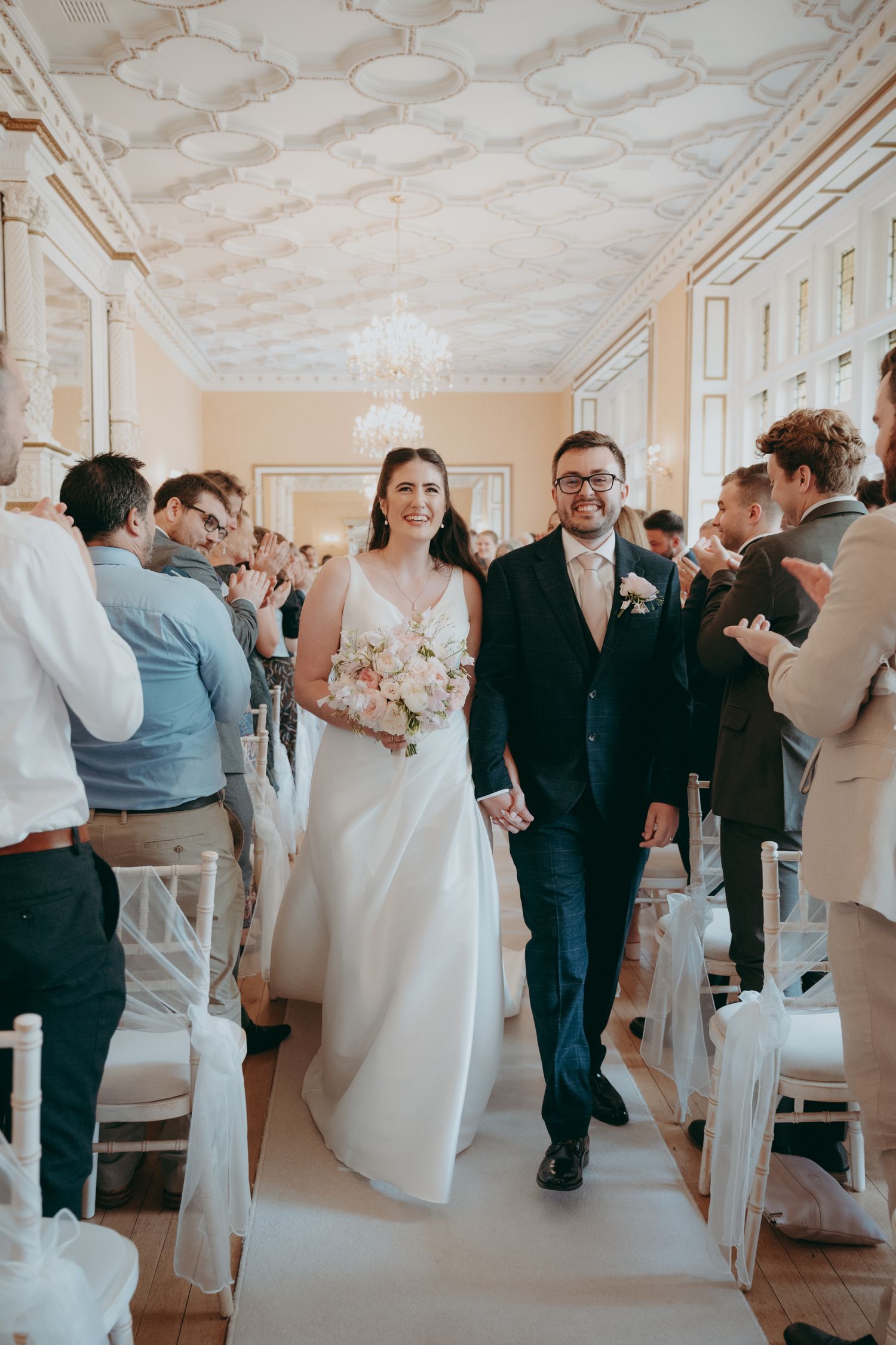 Bride and groom walk down the aisle smiling in an elegant venue with ornate ceiling and white chairs.