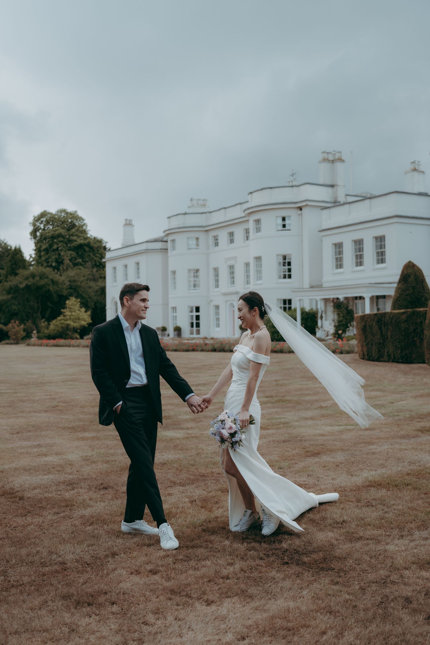 Bride and groom holding hands walking on lawn before grand white manor house on overcast wedding day.