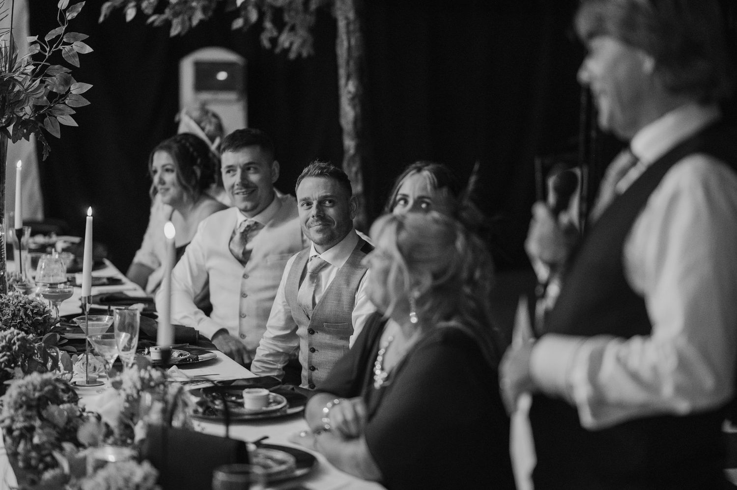 Black and white wedding reception photo of guests laughing at head table during heartfelt speech at elegant venue.