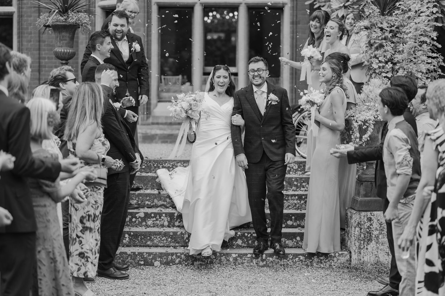 Black and white photo of a joyful newlywed couple walking through confetti shower surrounded by guests at elegant venue.