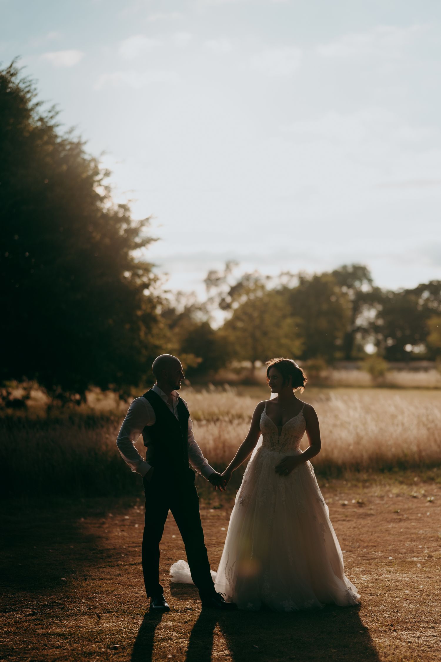 Bride and groom holding hands at golden hour in a field, bride in a flowing white gown with lace back detail.