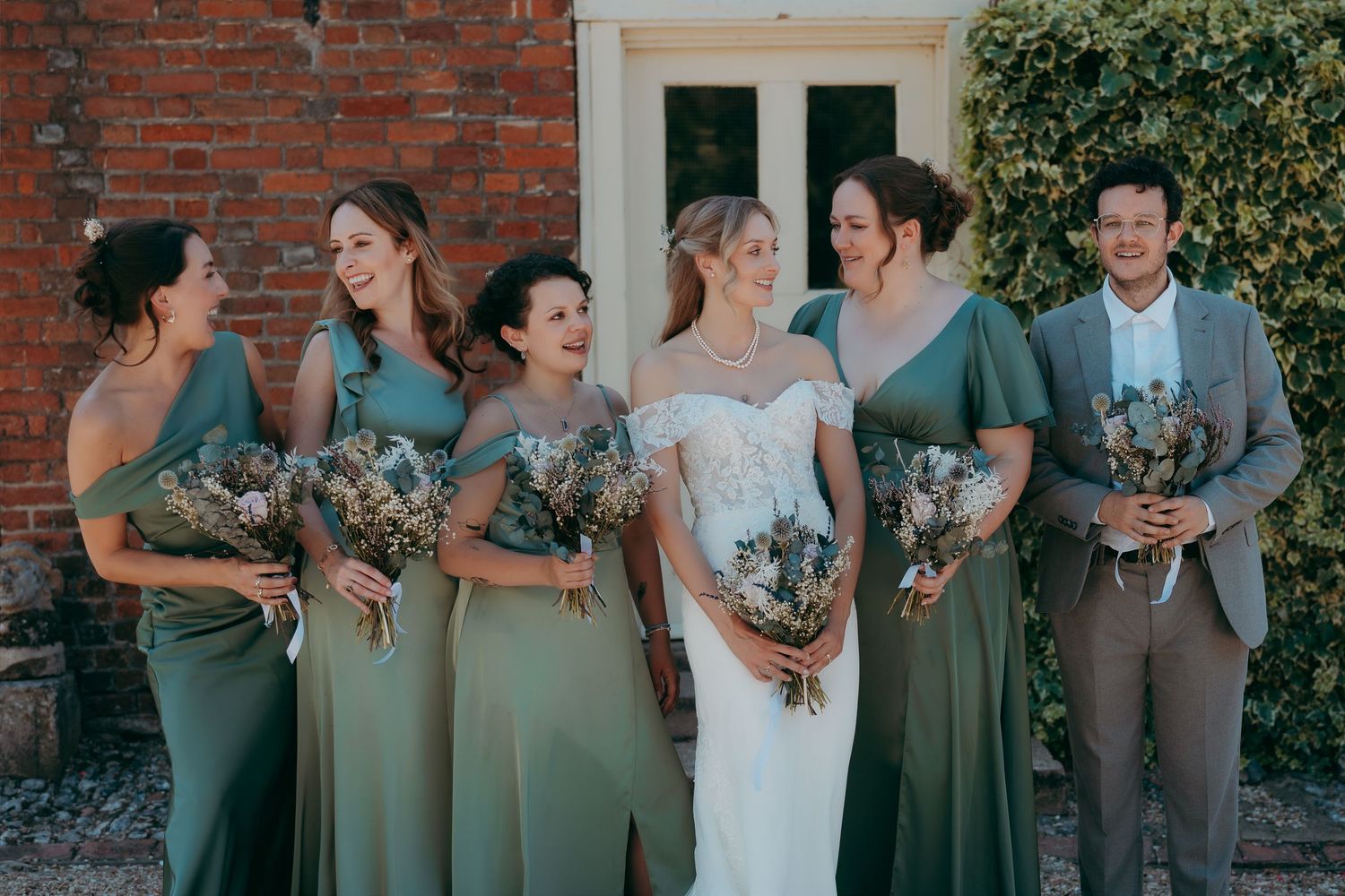 Bride in white lace gown with bridesmaids in sage green dresses holding dried bouquets outside a brick venue.
