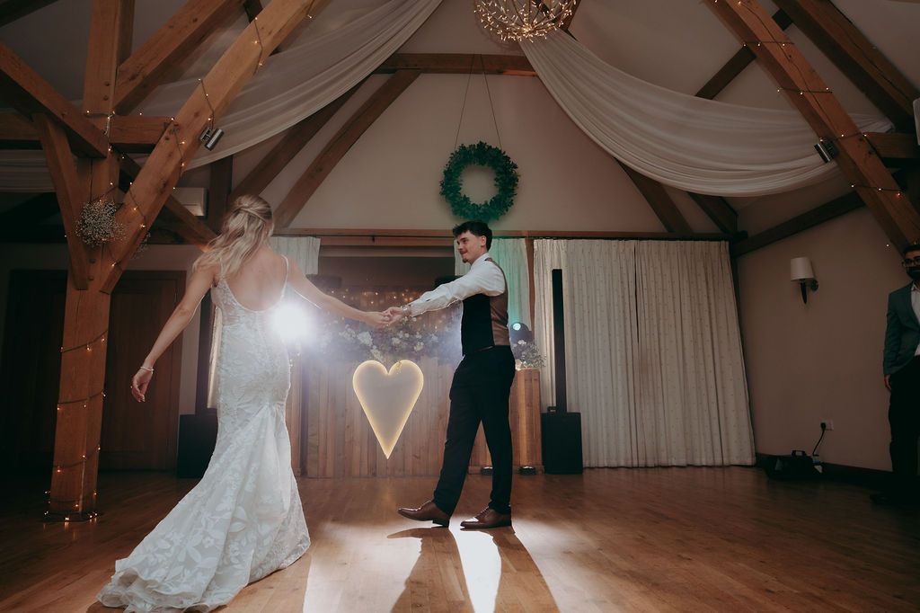 Bride in a lace wedding gown and groom in a vest share their first dance in a rustic barn venue with a glowing heart light and wreath.