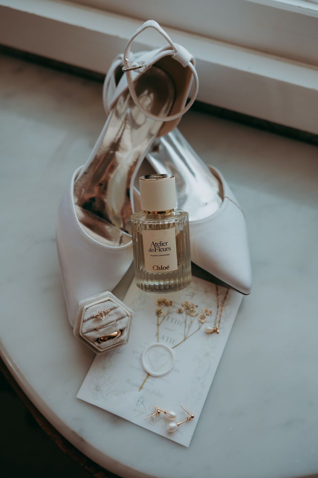 White pointed-toe bridal heels with ankle strap beside Chloé perfume, ring box, and delicate gold jewelry on a marble surface.